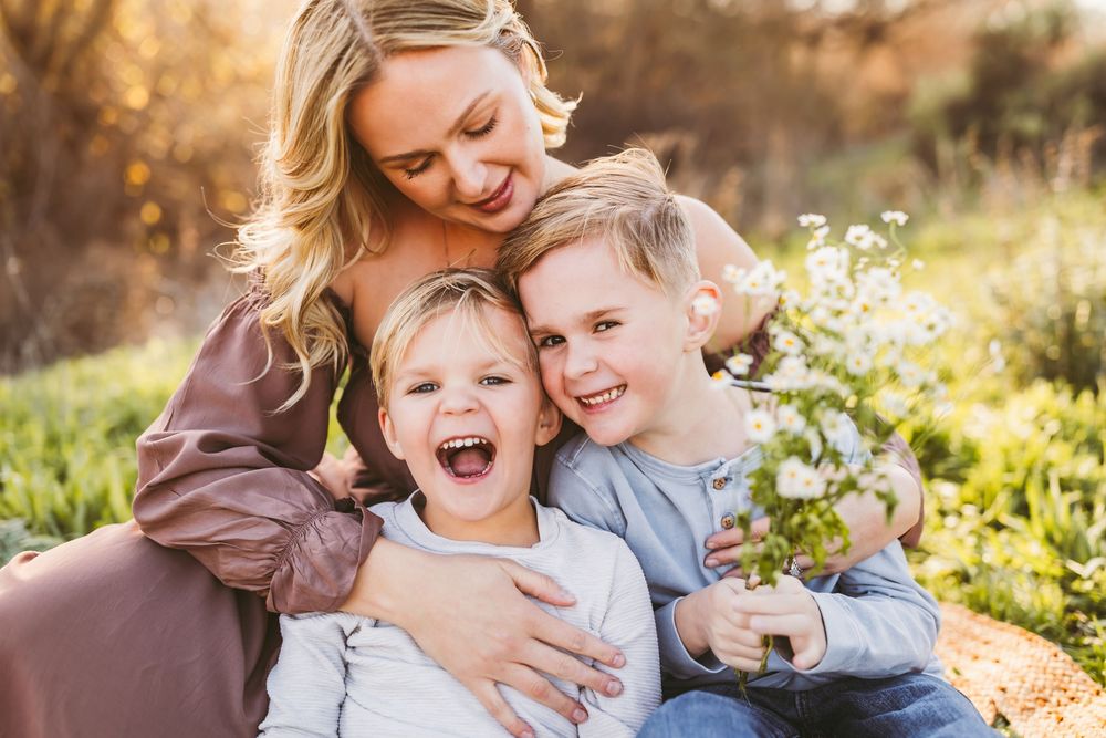 A smiling mother hugs her two laughing children outdoors in a sunny field with heidi grace photography