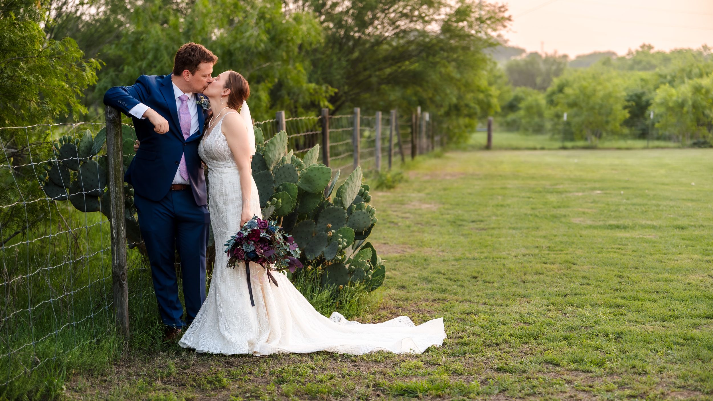 Romantic wedding portrait of a bride and groom kissing by a rustic fence in a lush, green Central Texas field.