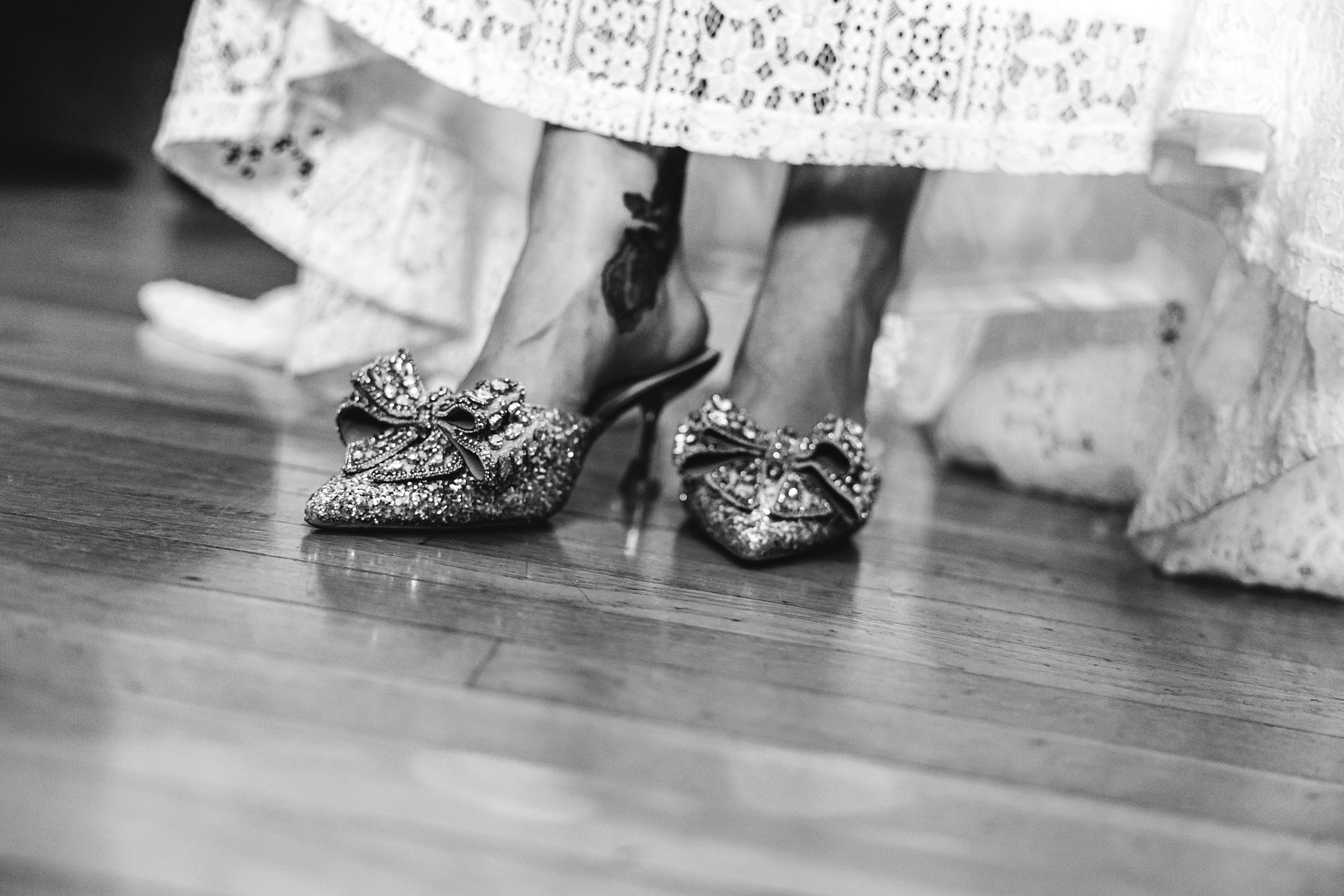 Black and white detail shot of a bride's sparkling pointed-toe wedding shoes and the lace hem of a wedding dress.