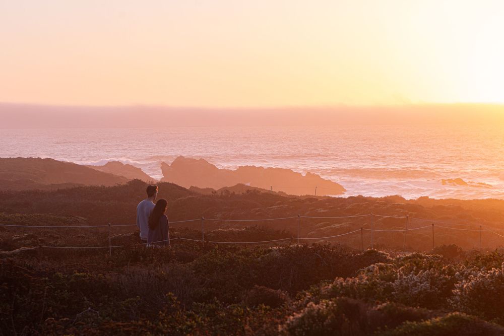 Two people watch a stunning golden sunset over the ocean from a scenic coastal overlook surrounded by low shrubs.