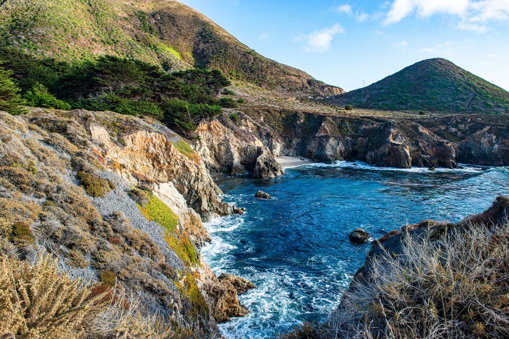 Rocky coastal cliffs overlook a vivid blue cove surrounded by green hills in Big Sur, California.
