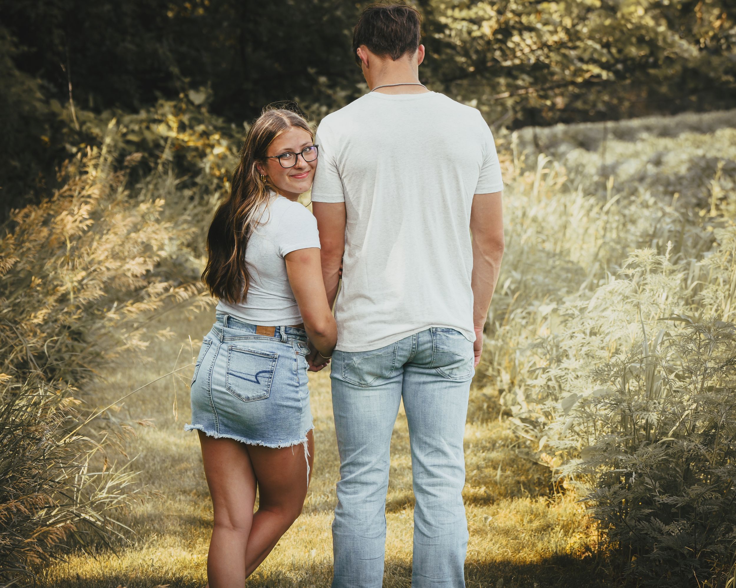 A couple holds hands while walking through a sunlit meadow, the woman wearing glasses and a denim skirt looks back.