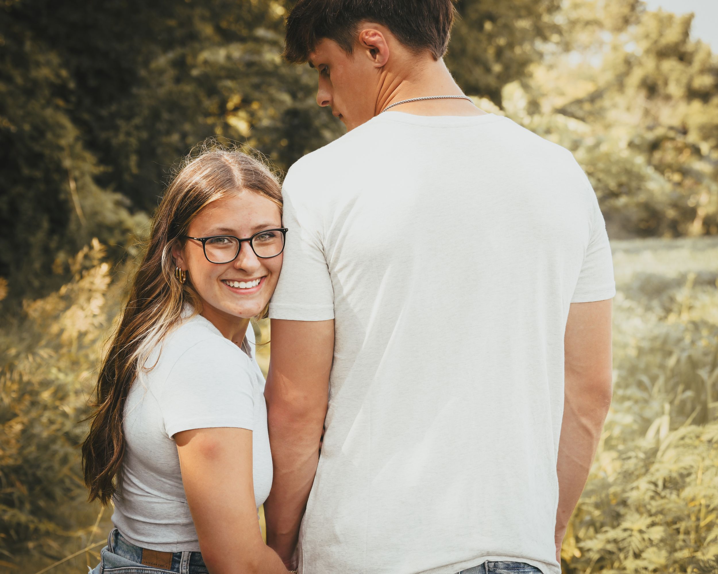 A smiling young woman with glasses in a white top holds hands with a young man outdoors in a sunny, green setting.
