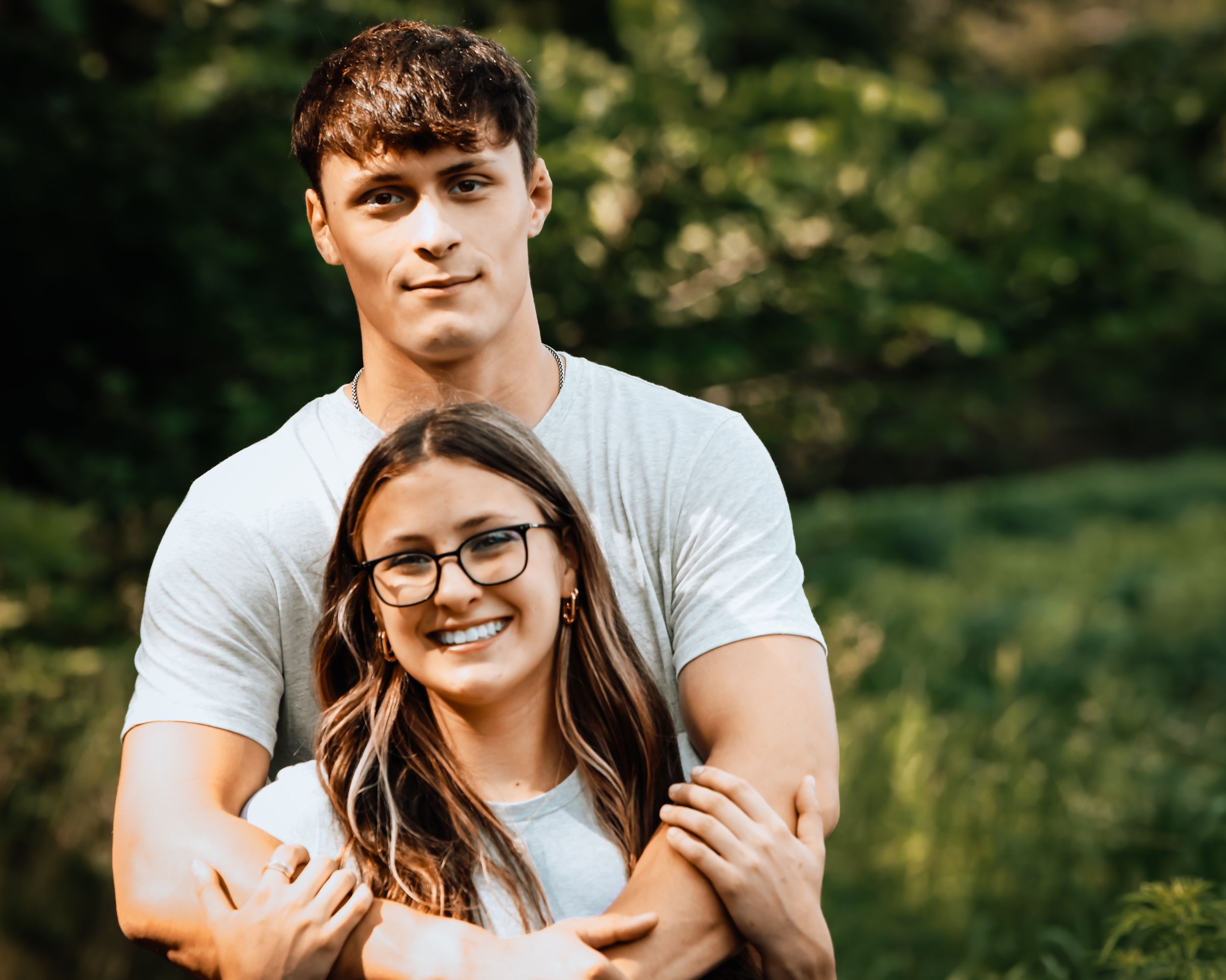 Young couple embracing outdoors, man hugging woman from behind, both smiling, wearing white shirts with green trees background.