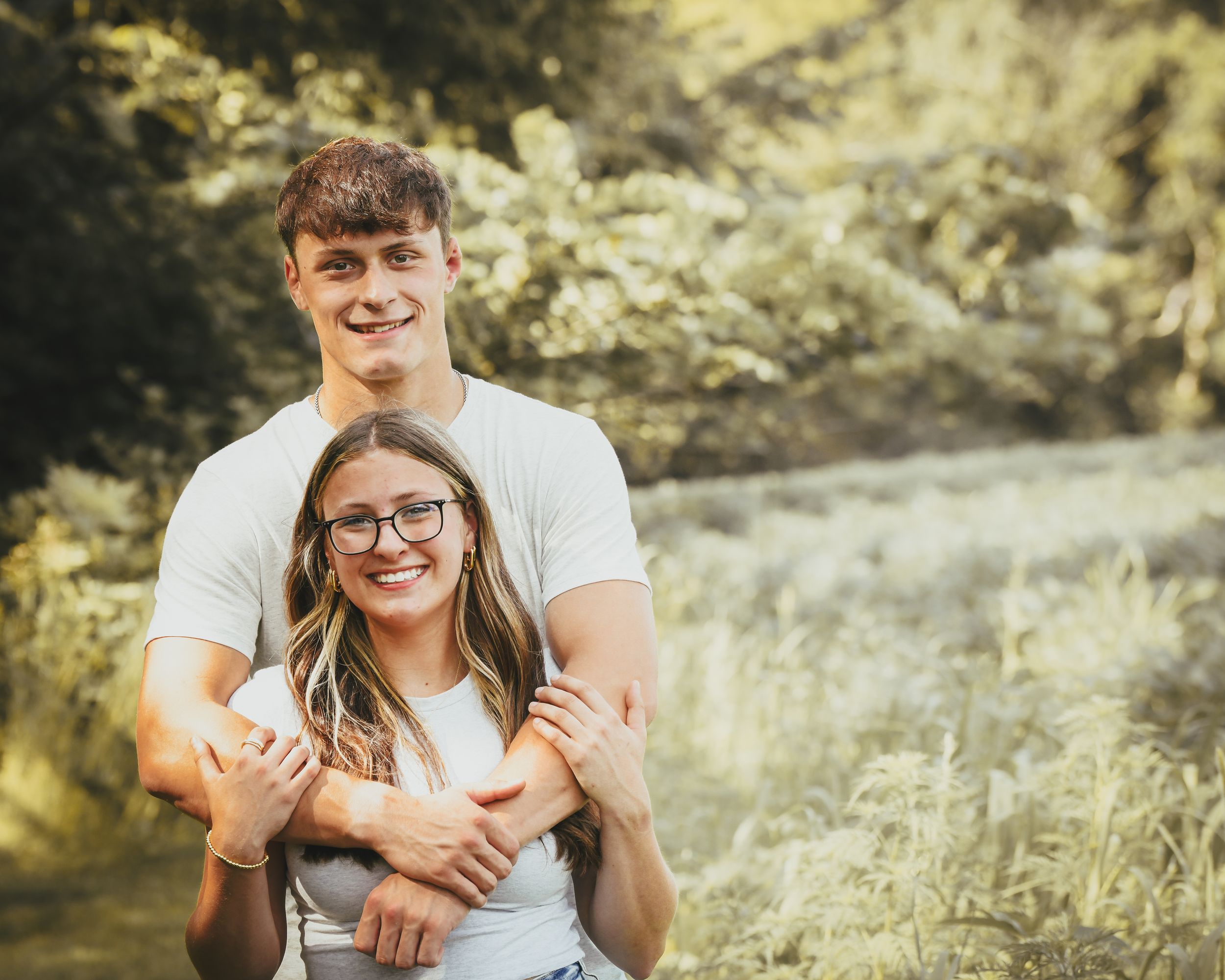 Young couple embracing outdoors in a lush green setting, smiling warmly for a portrait photo.
