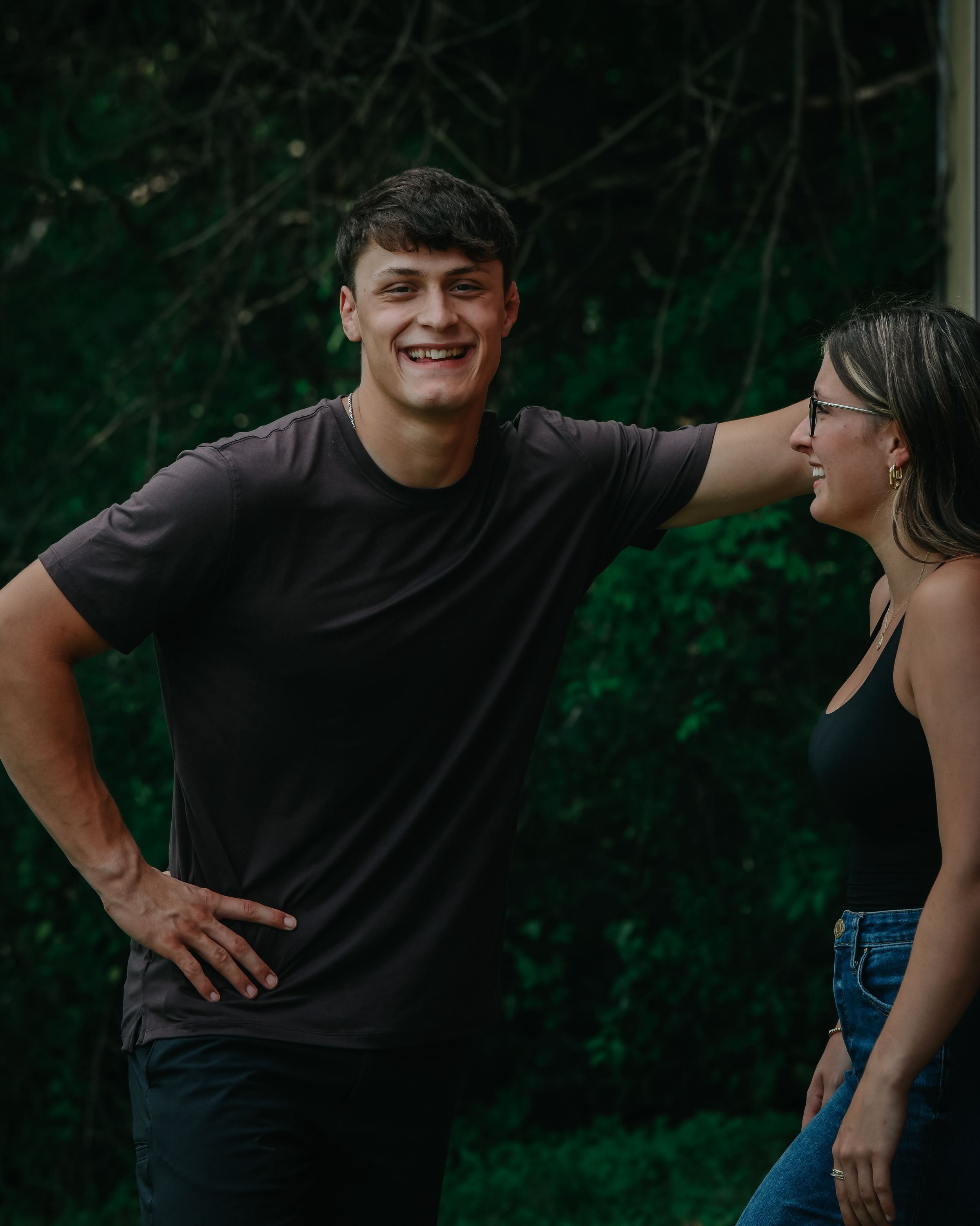 A laughing young man in a black t-shirt stands beside a smiling woman in a dark top outdoors.