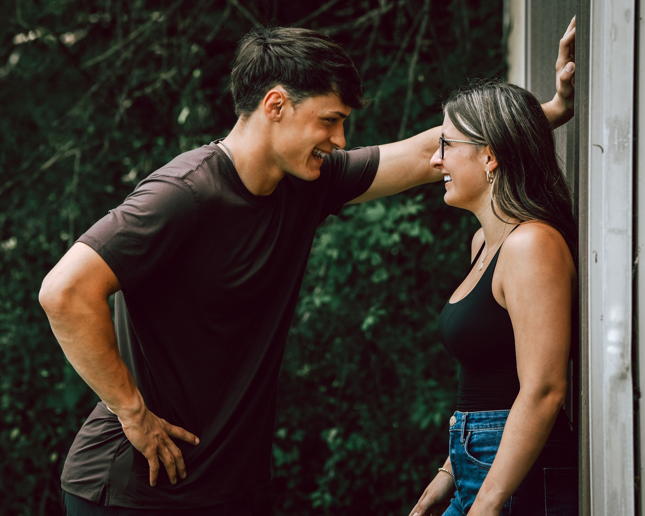 Young couple in black outfits sharing a romantic moment outdoors, man leaning against wall smiling at woman.