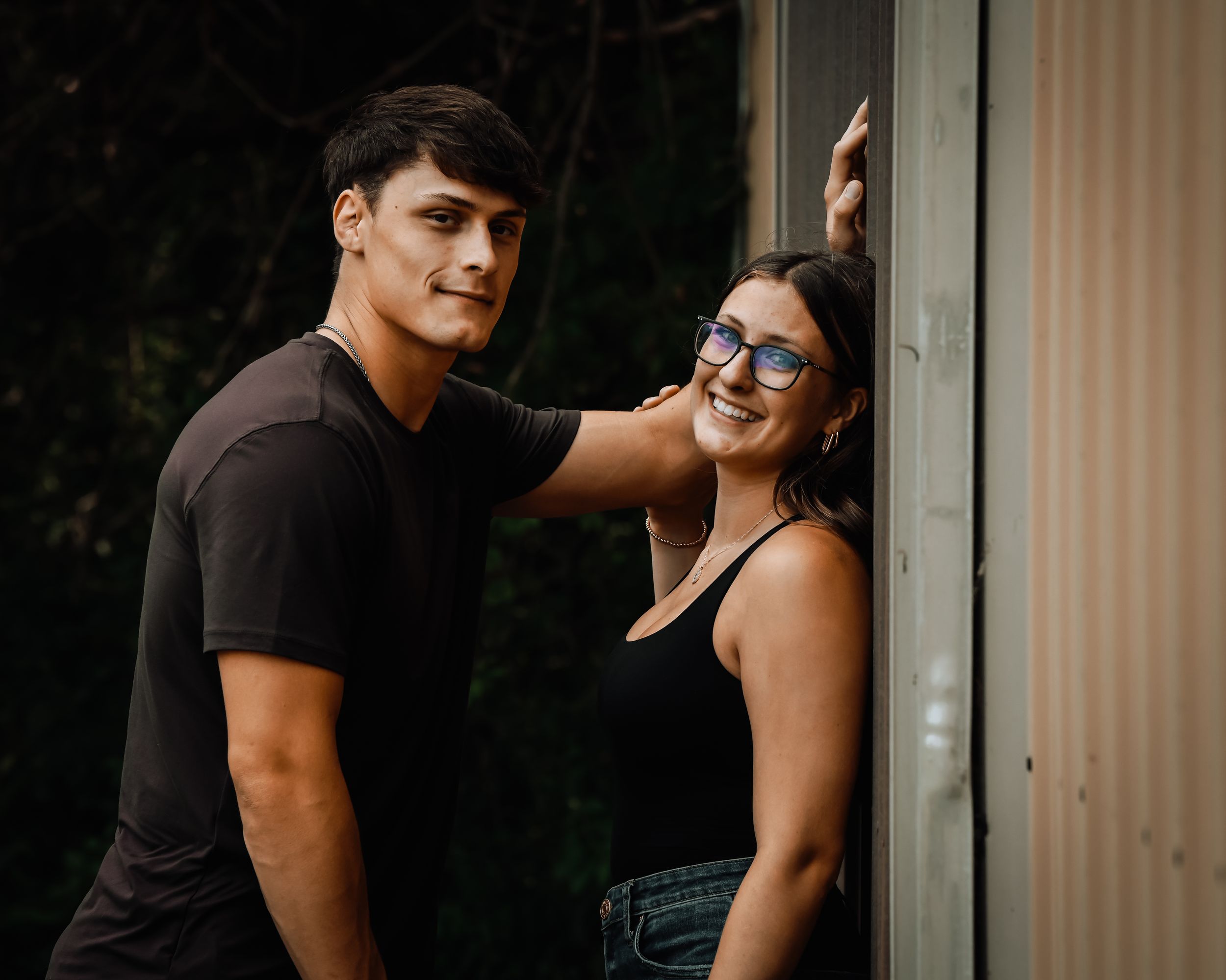 A young couple poses outdoors, man in black tee with hand on wall, woman in tank top and glasses smiling warmly.