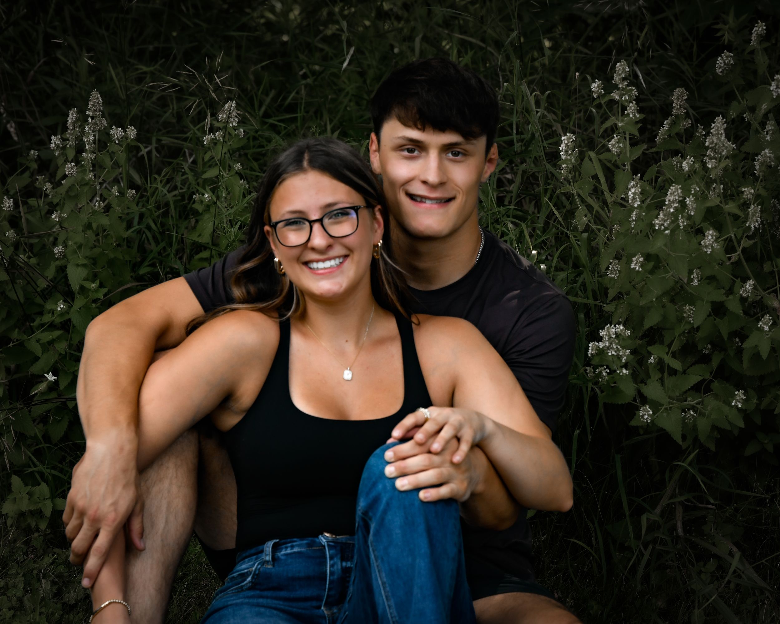 Smiling couple sitting together in front of lush green foliage, woman wearing glasses and black top.