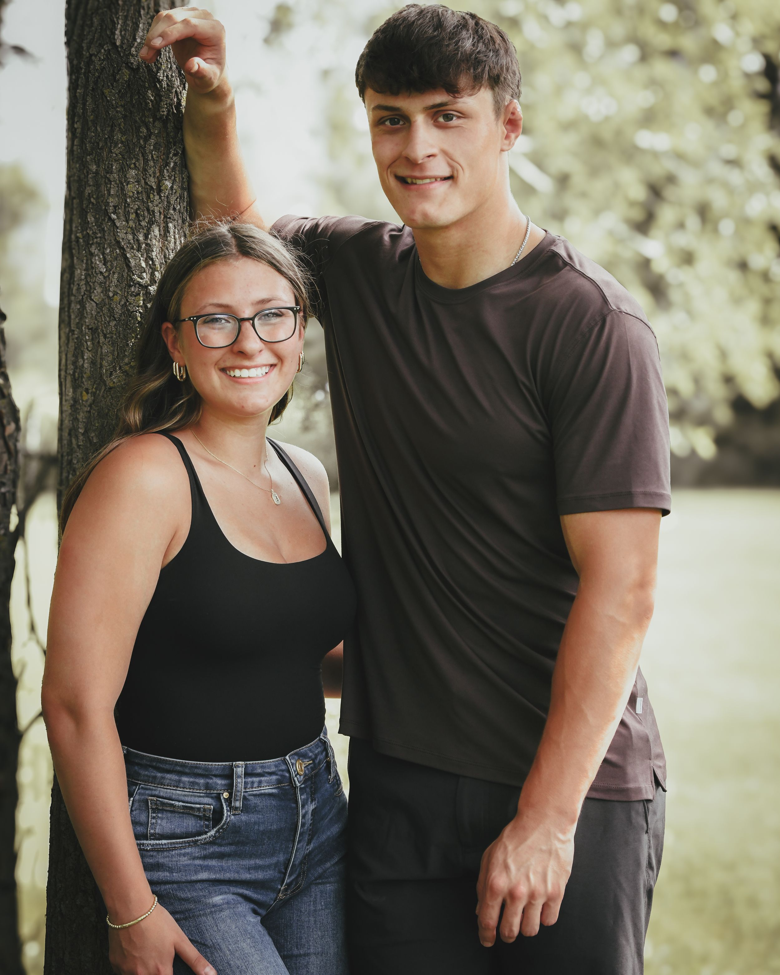 A young couple smiles together outdoors, leaning against a tree in a lush green park setting.
