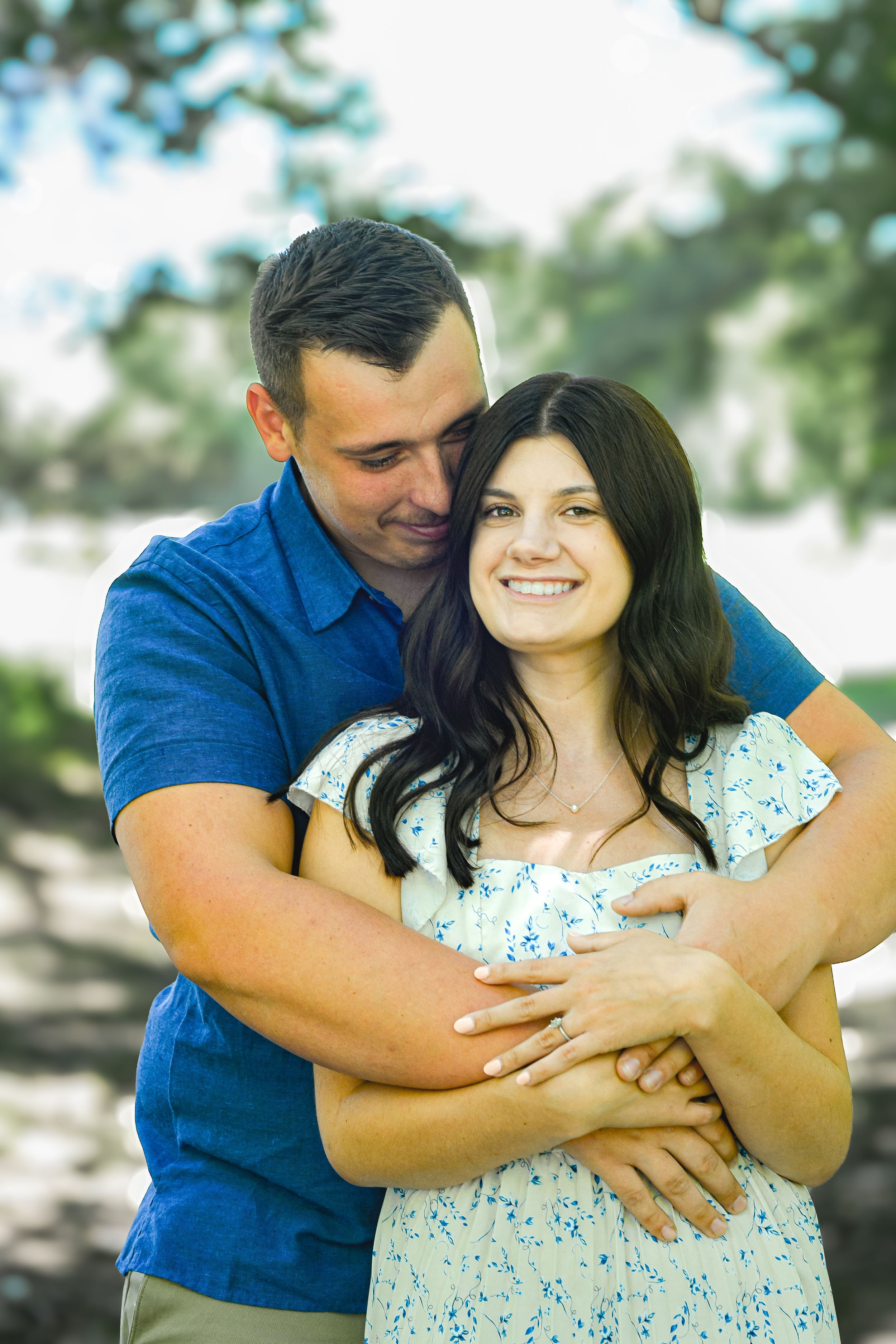 A couple embraces outdoors, woman smiling in floral dress as man in blue shirt wraps arms around her.