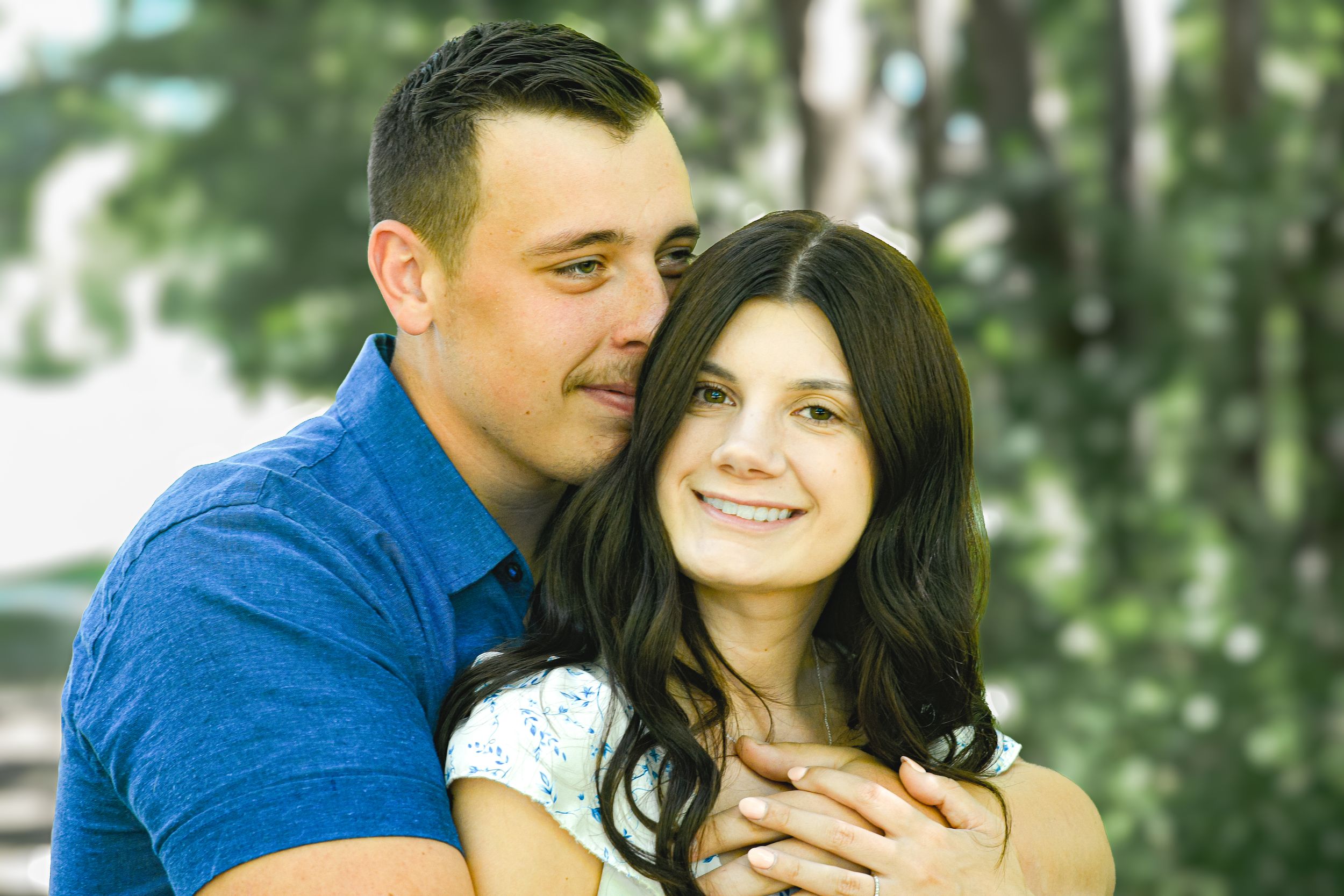 Happy couple embracing outdoors, smiling woman with dark hair and man in blue shirt, with green trees in background.