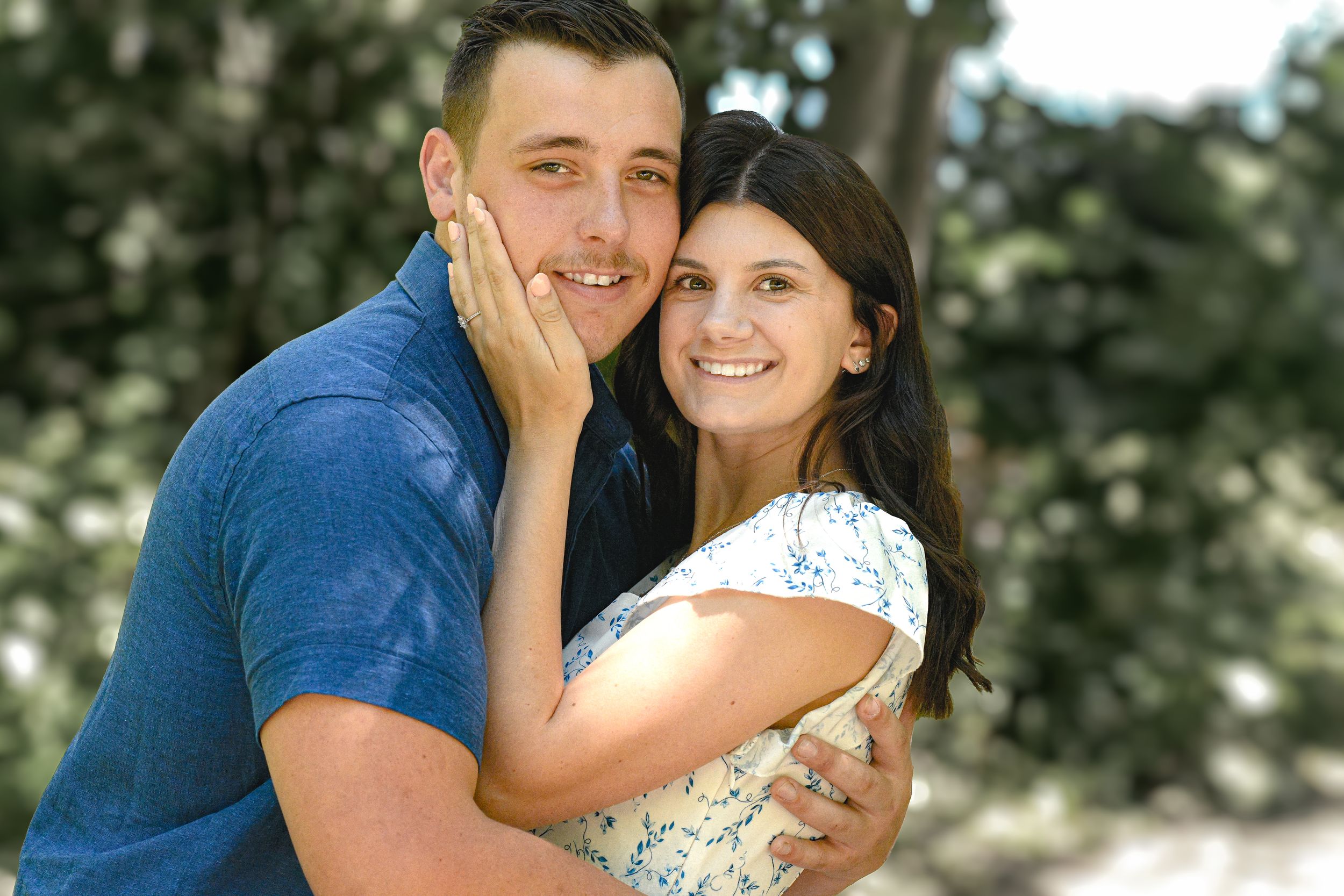 Happy couple embracing outdoors, smiling woman in floral dress and man in blue shirt posing together in a sunny park.