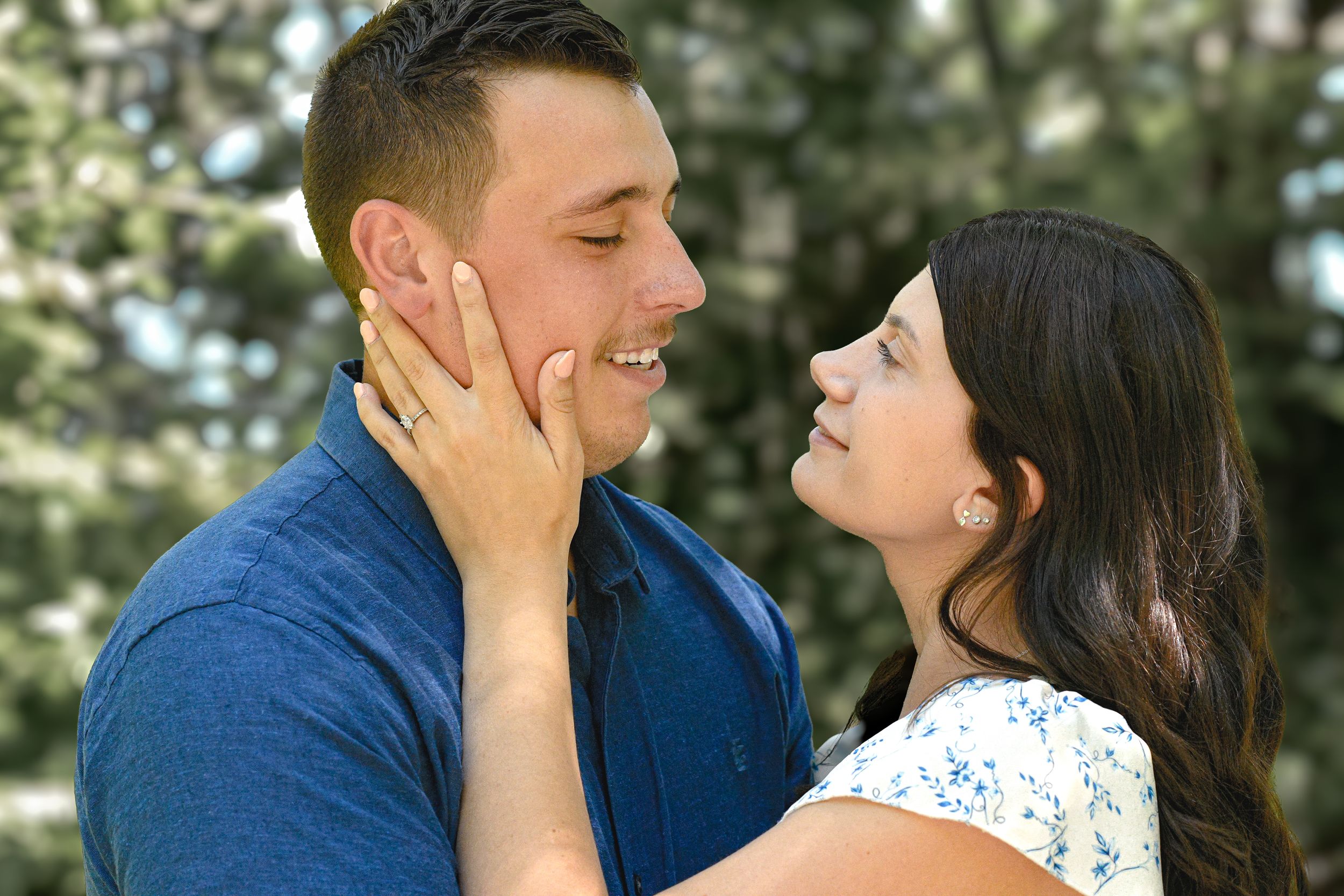 A smiling couple faces each other outdoors, woman gently holding mans face in a romantic moment.