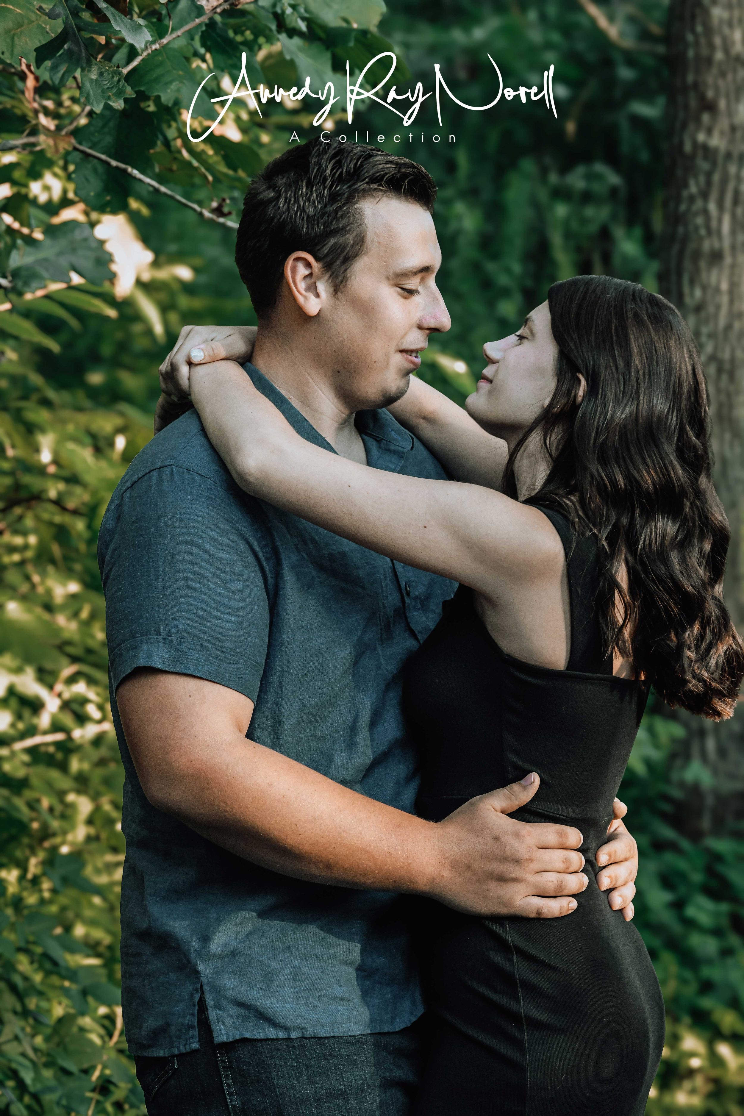 A couple embraces tenderly outdoors, the woman in black dress gazing up at the man amid lush green foliage.