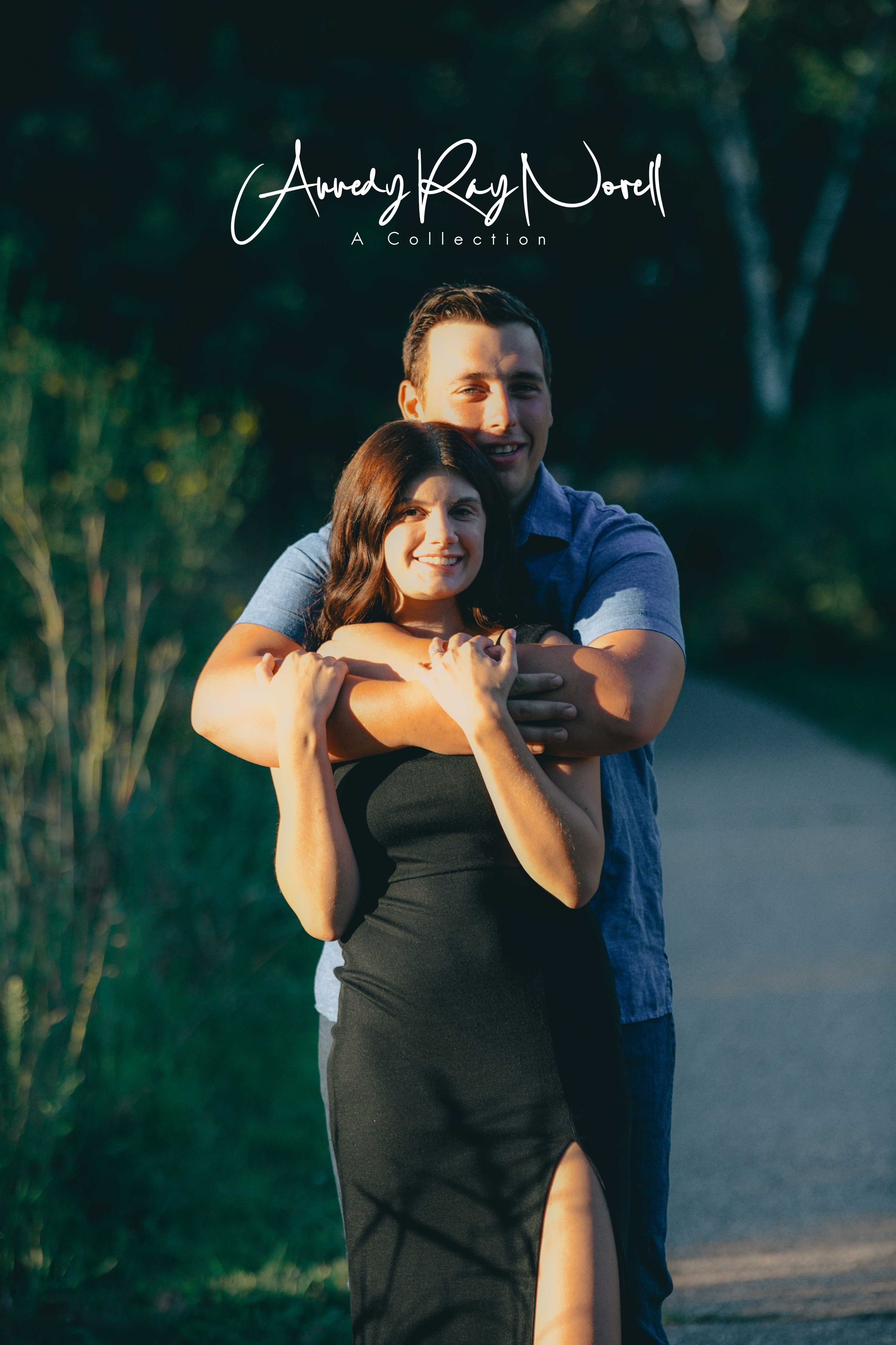 A couple embraces on a garden path at golden hour, the man hugging the woman from behind as she smiles warmly.