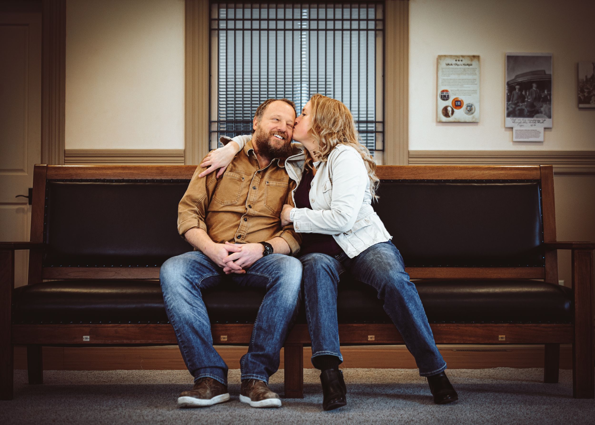 Couple sitting on a wooden bench sharing a tender moment, woman kissing man's cheek in a cozy indoor setting.