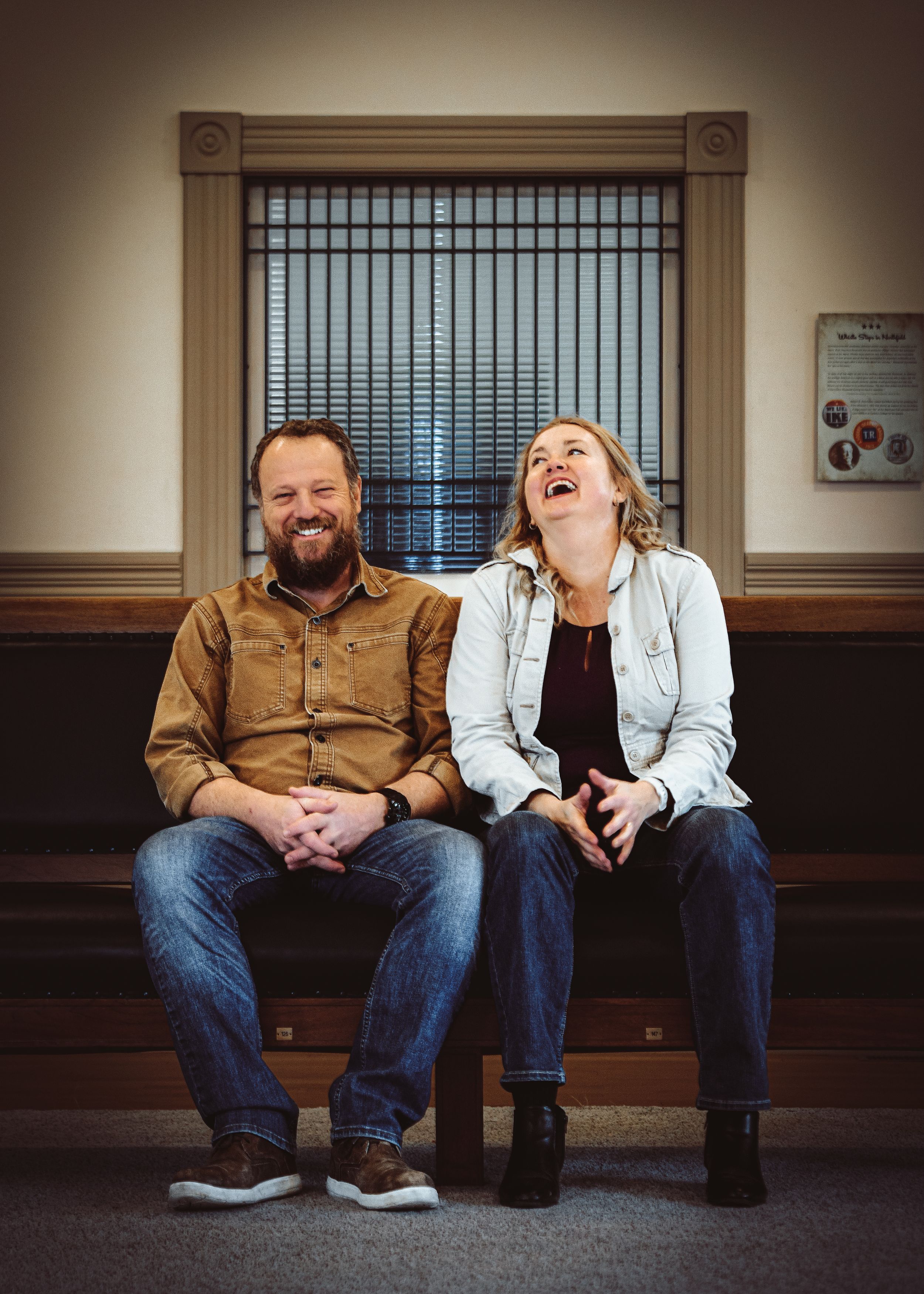 Two adults laughing joyfully while seated on a bench indoors, wearing casual denim outfits in a warmly lit room.