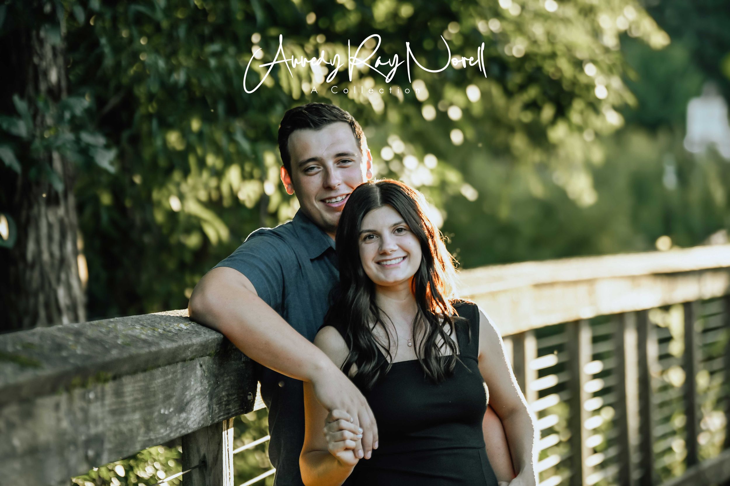 Smiling couple posing on a wooden bridge surrounded by lush green trees in a park setting.