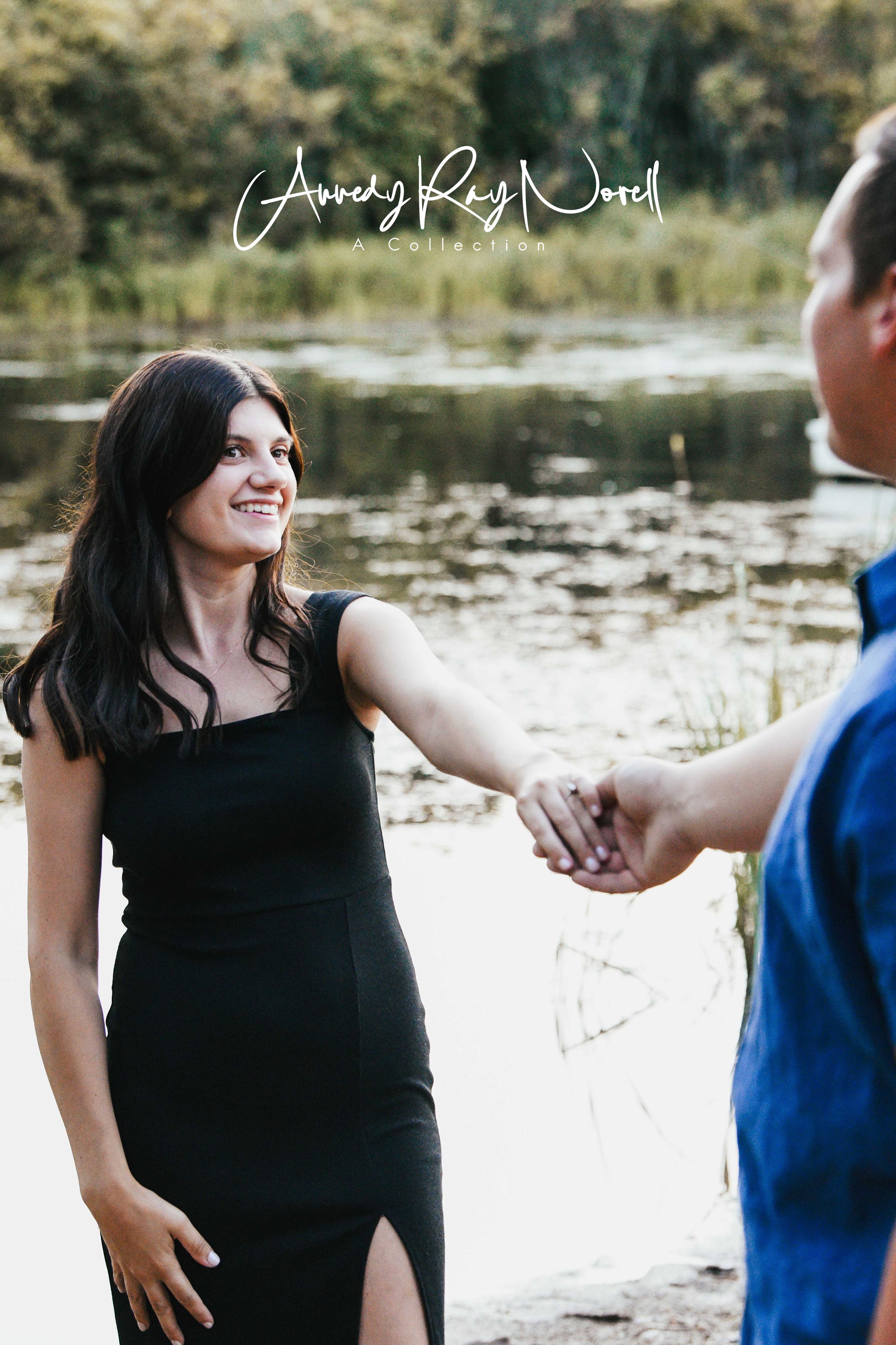 Couple holding hands near a serene pond, woman in black dress smiling, outdoor engagement photo session.