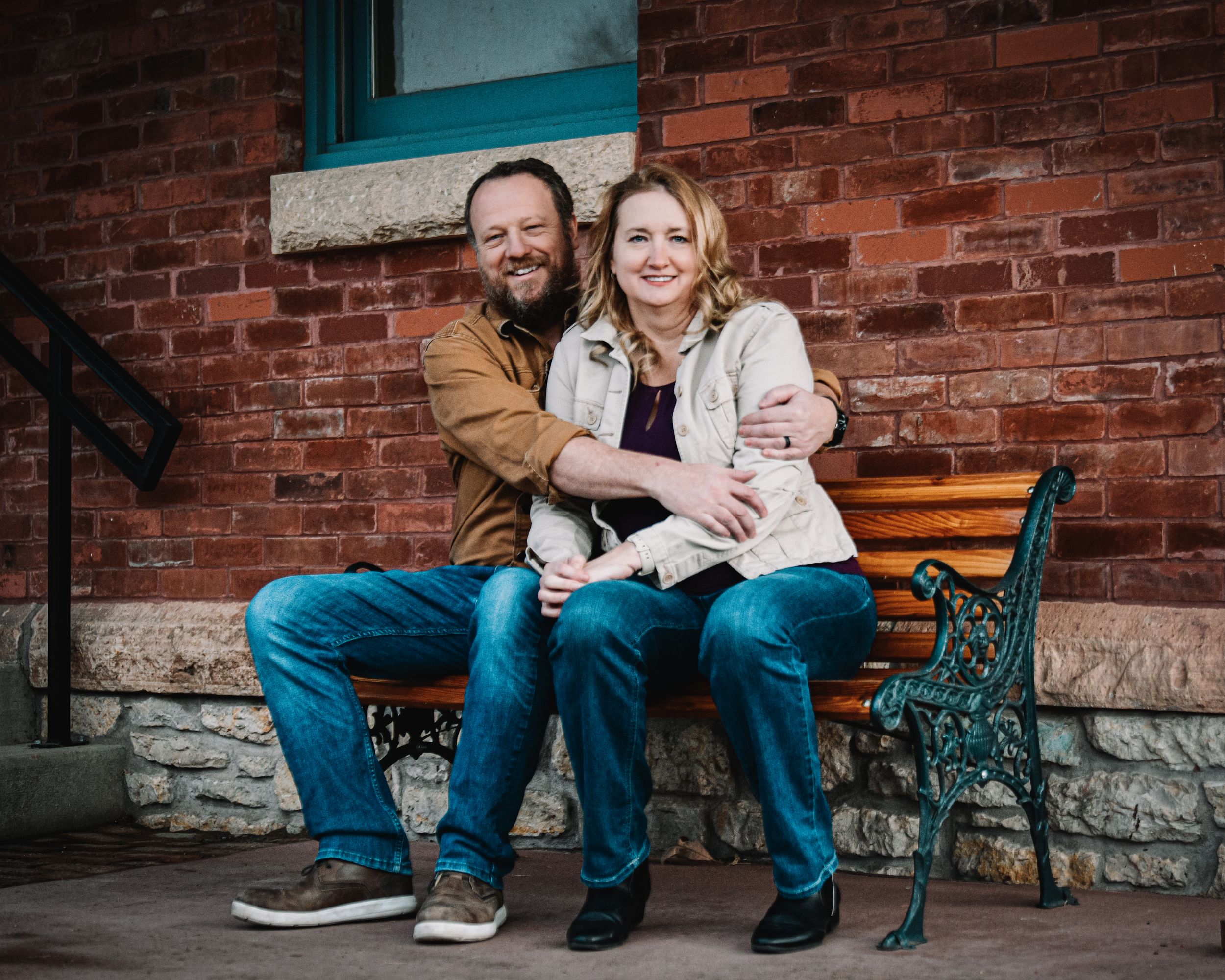 Couple sitting together on a wooden bench in front of a brick building, smiling warmly in casual attire.