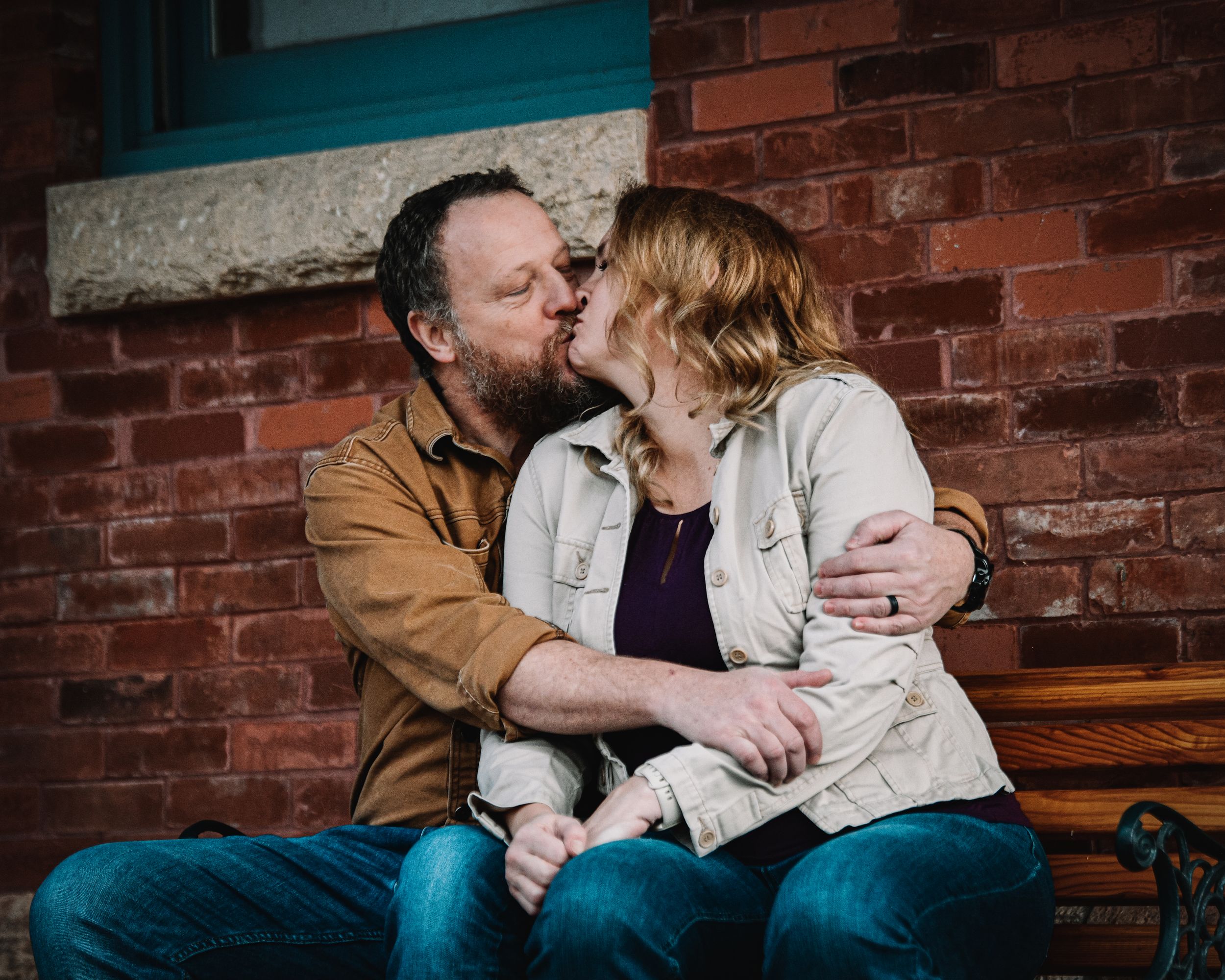 Couple sharing a romantic kiss on a bench against a red brick wall, man in brown jacket embracing woman in beige coat.