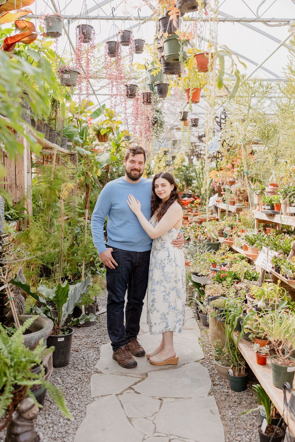 Couple on a stone path in a bright greenhouse with green plants and warm light; he wears a blue sweater, she a white floral dress.