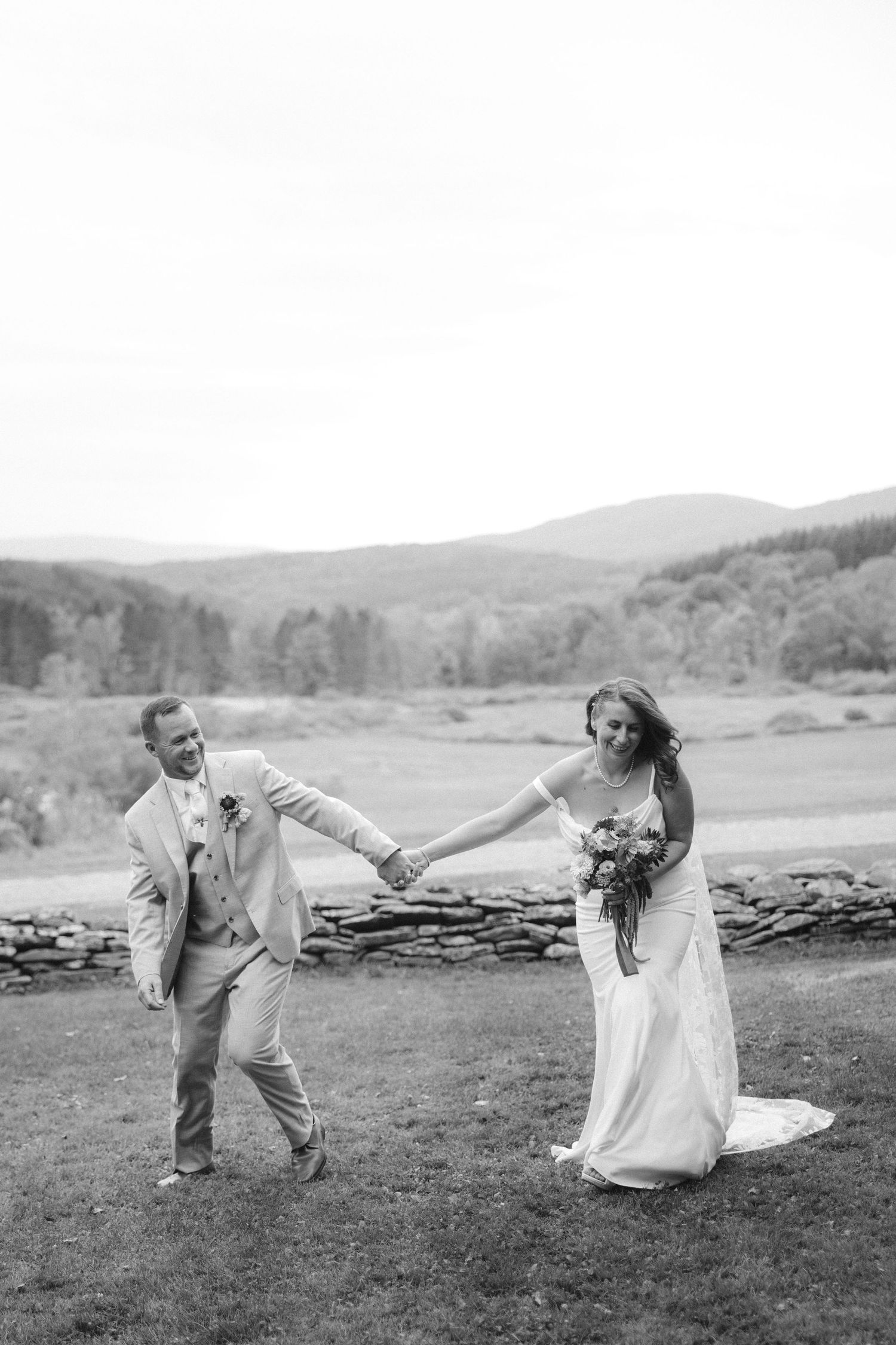Black and white photo of a bride and groom holding hands, dancing in a field with rolling hills in the background.