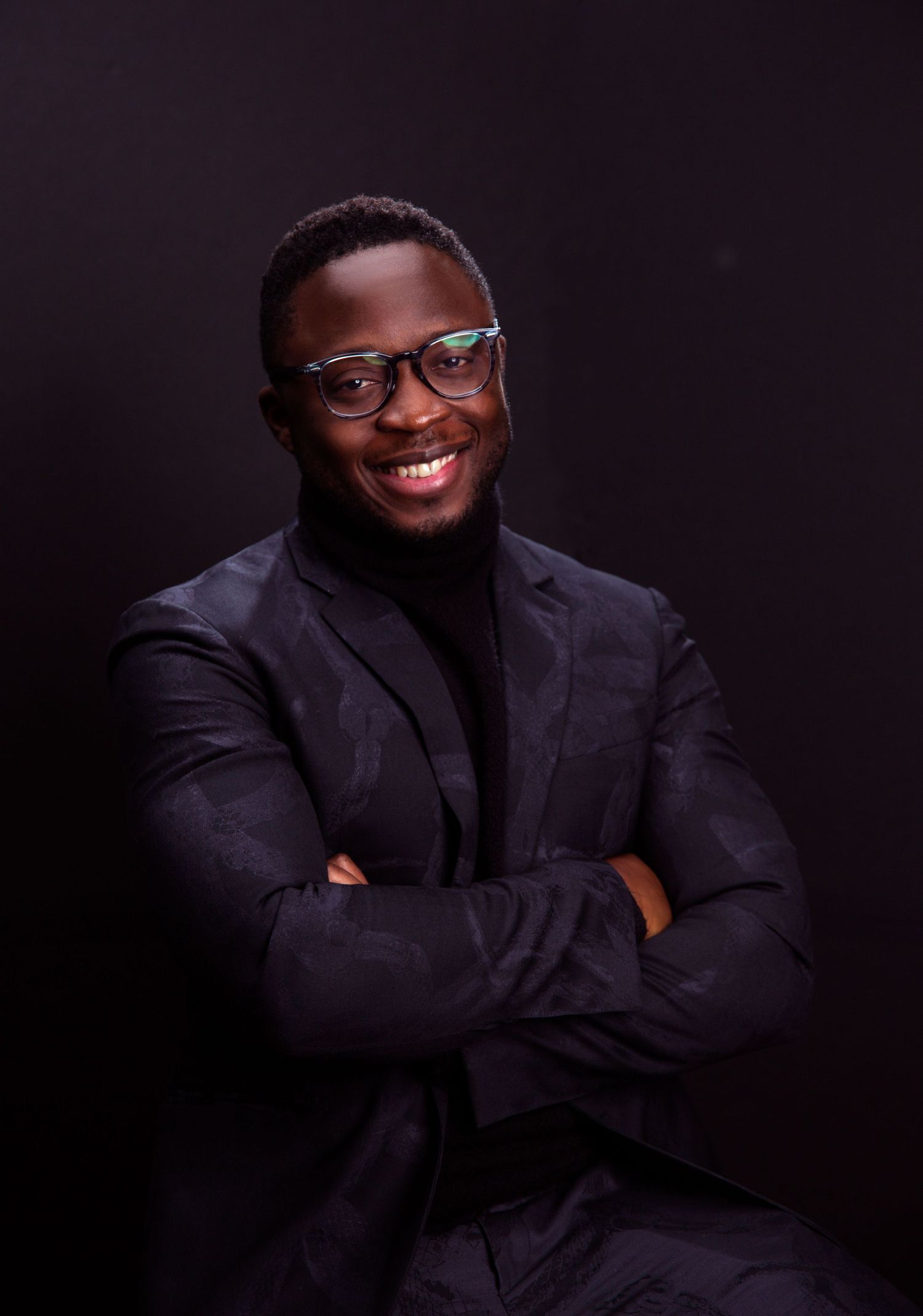 Confident young man in black suit and glasses smiling with arms crossed against dark background.