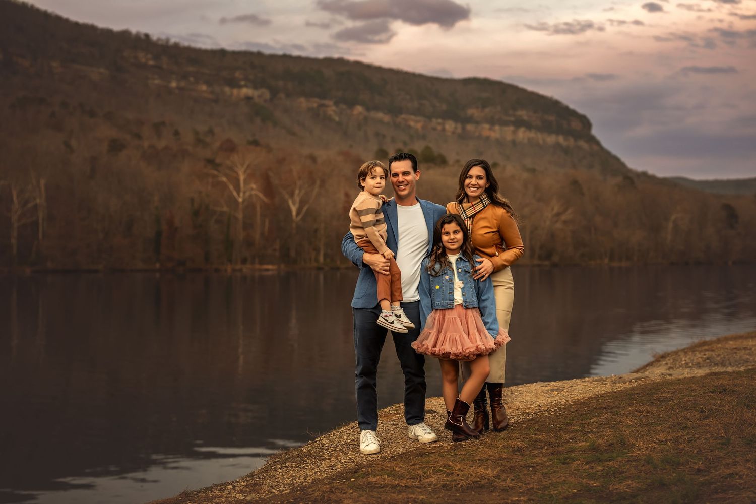 Family of four posing by a lake with autumn hills at dusk.
