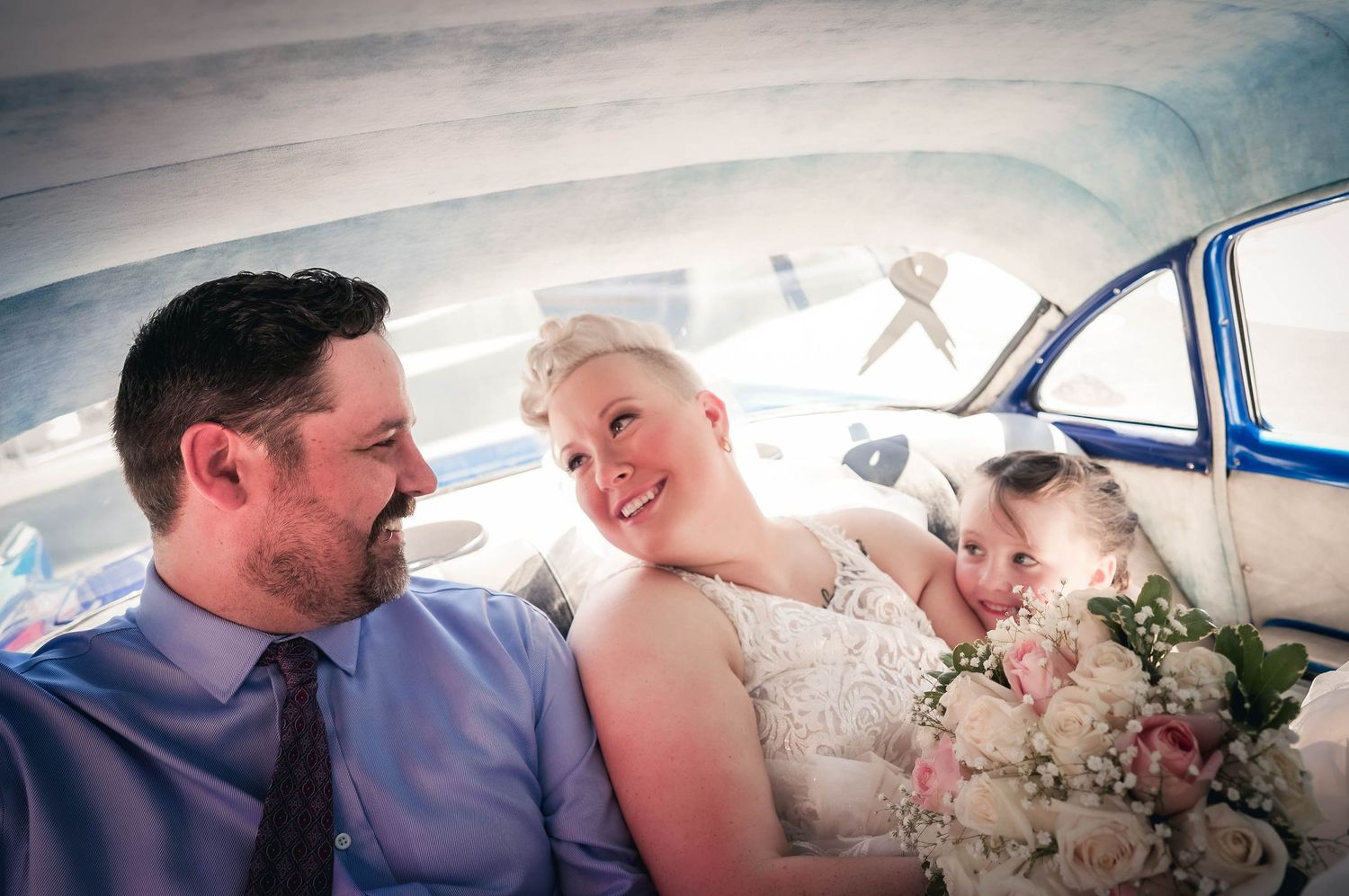 Newly married couple smiling at each other with a cute flower girl peeking at them, by the best local photographer