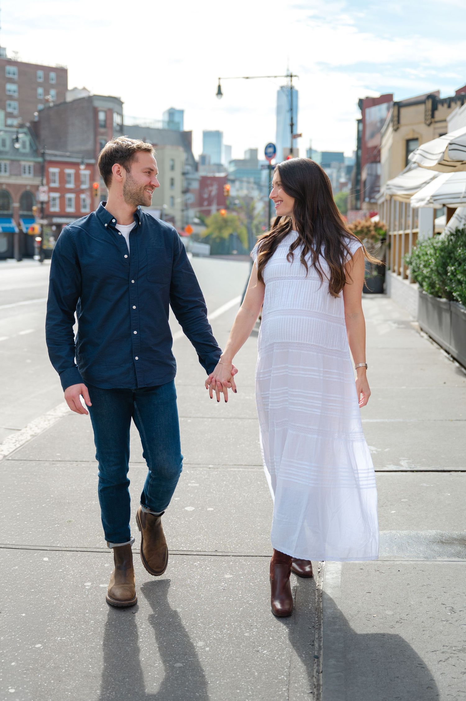 Couple holding hands walking on a city sidewalk, man in navy shirt and jeans, woman in white maxi dress and boots.