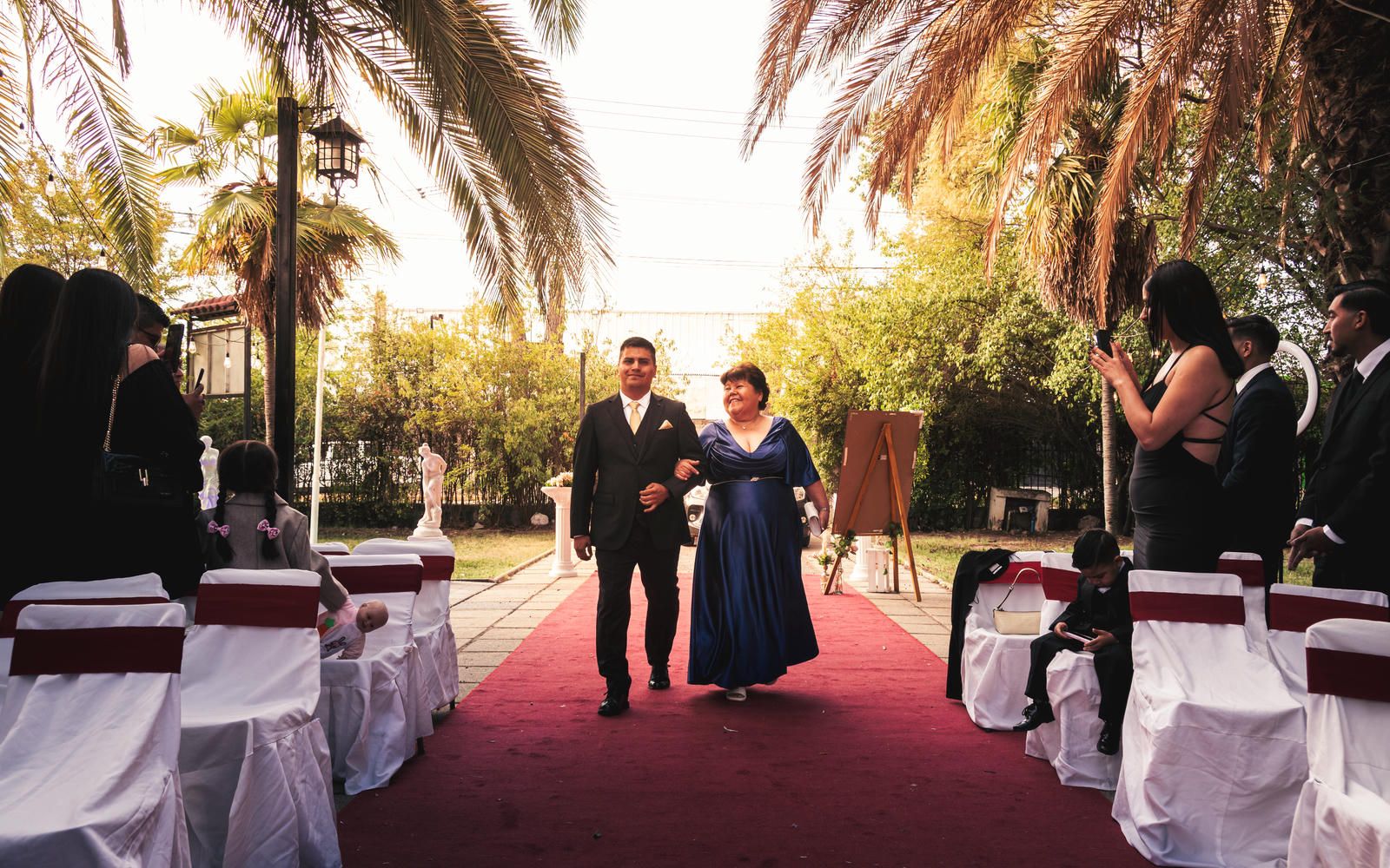 Couple walks down red carpet aisle at outdoor wedding ceremony surrounded by palm trees and white chairs.