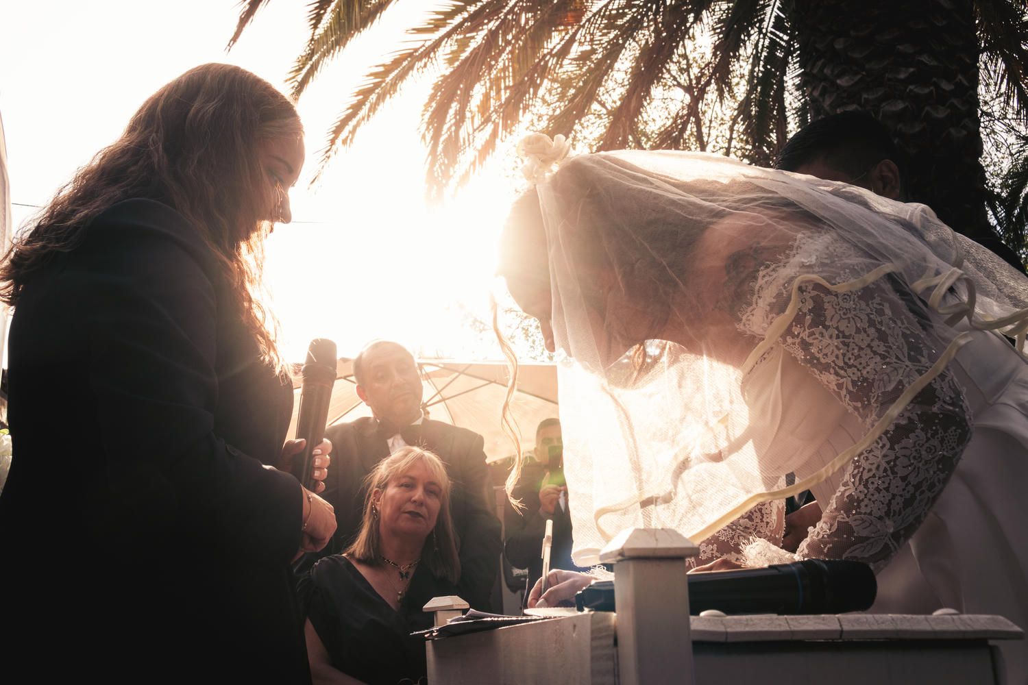 Bride in lace wedding dress bowing during outdoor ceremony with golden sunset light through palm trees.