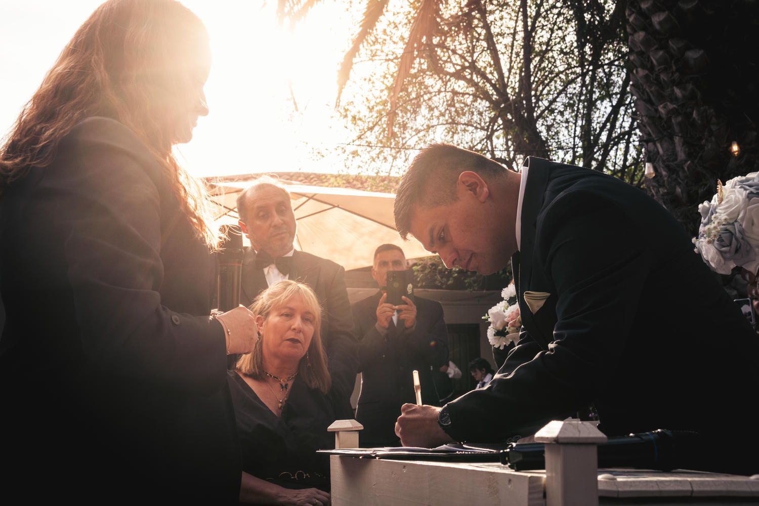 Groom in suit signing wedding documents outdoors at golden hour surrounded by guests.