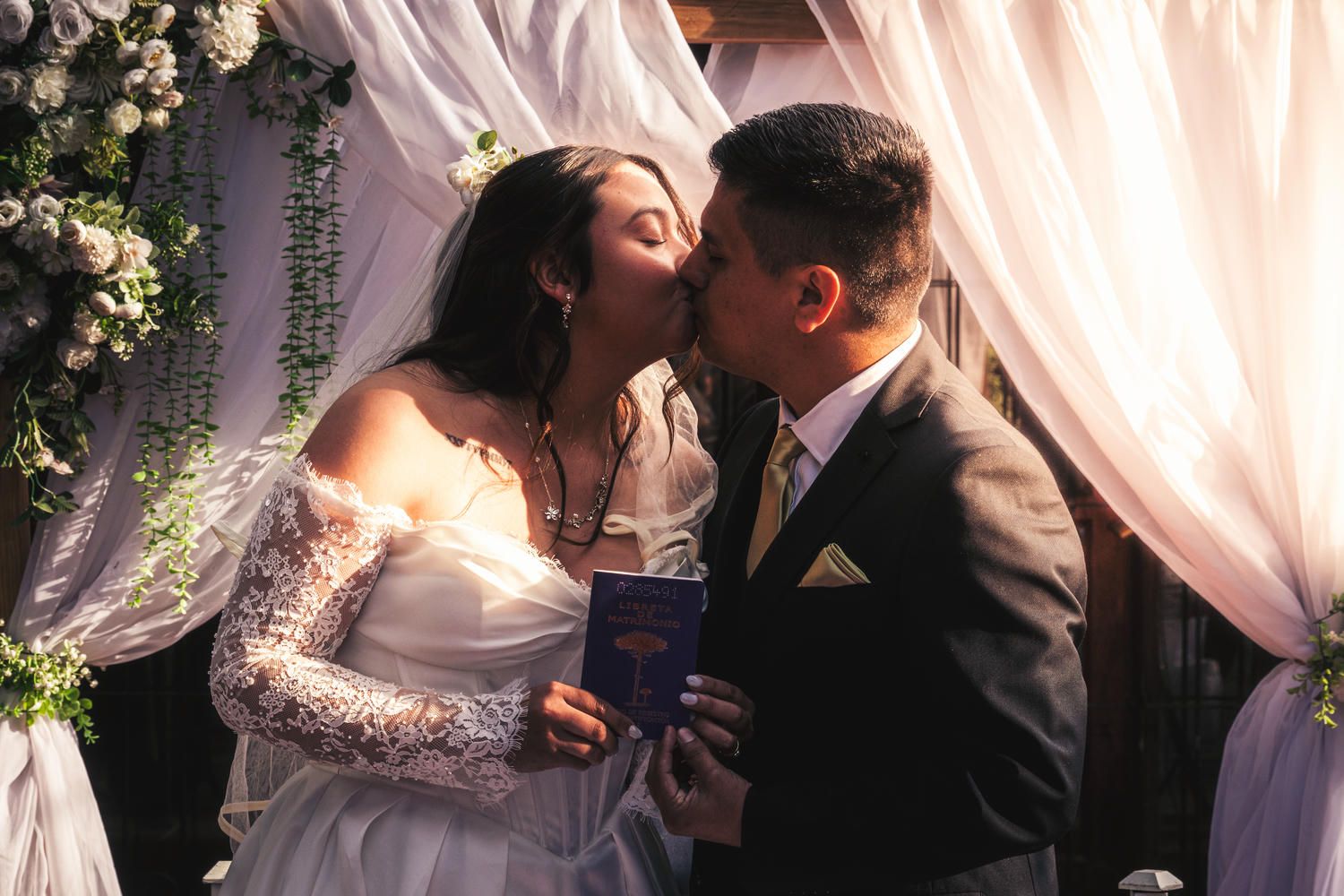 Bride and groom share a kiss at the altar during their wedding ceremony beneath a floral arch with white draping.