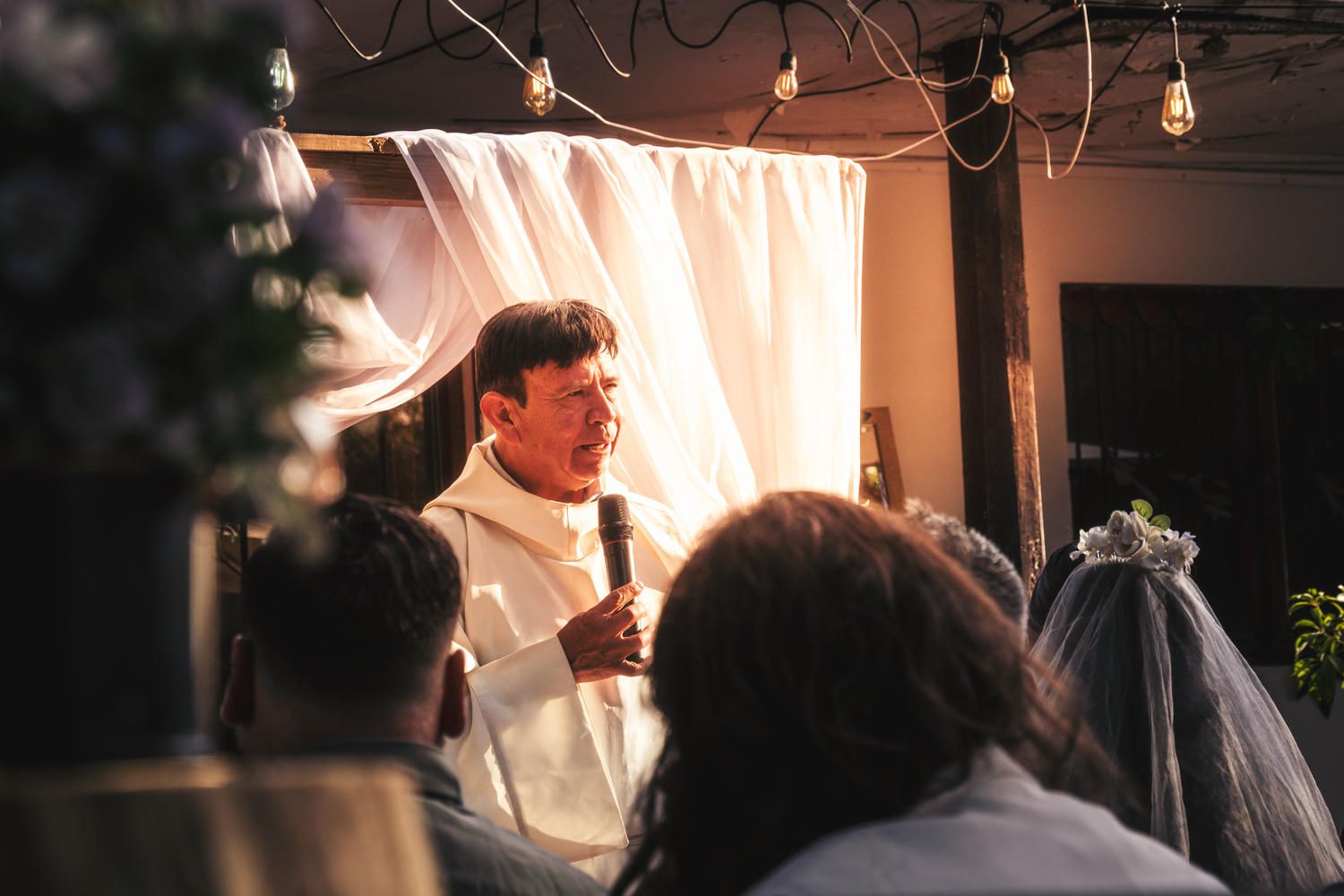 A priest in white robes officiates a wedding ceremony indoors with soft string lights and draped white fabric backdrop.