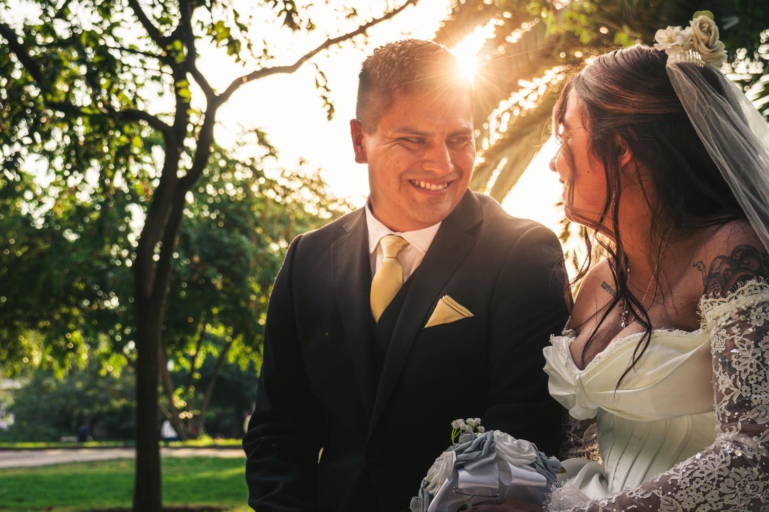 A smiling bride and groom share a romantic moment outdoors with golden sunlight filtering through green trees.