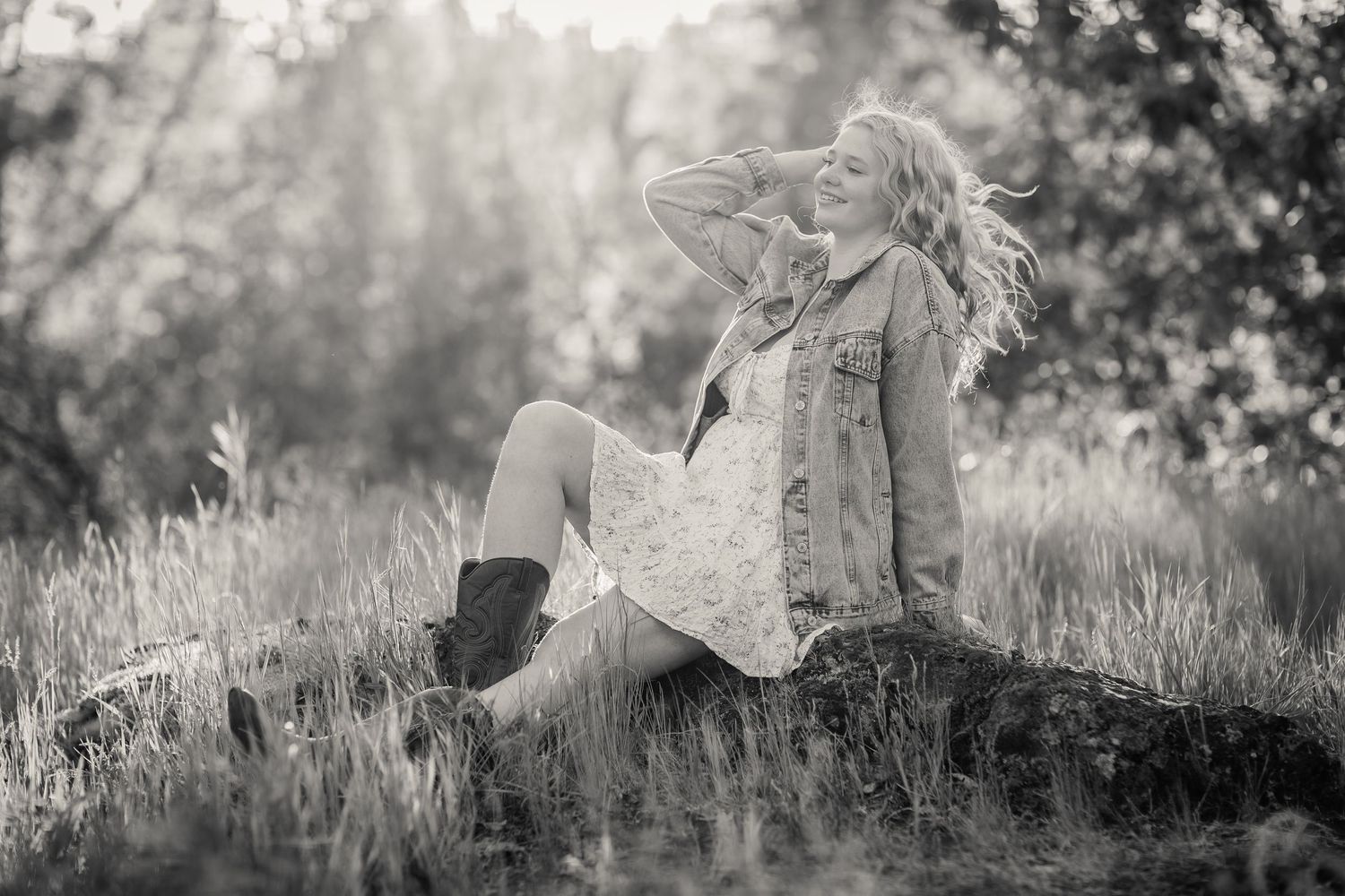 Black and white photo of a smiling blonde woman sitting on a rock in a field, wearing a denim jacket and cowboy boots.