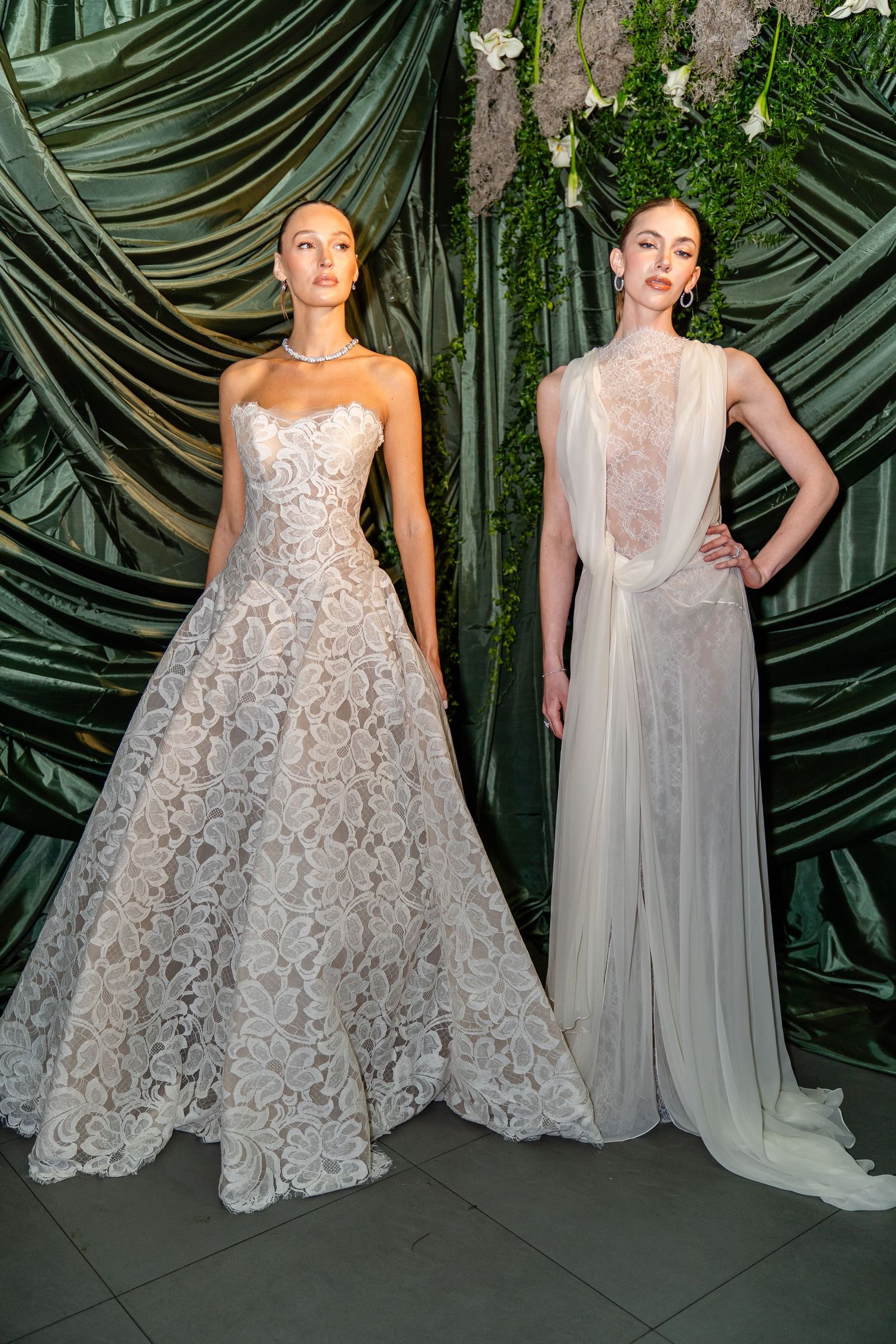 Two models wearing elegant bridal gowns, one lace A-line and one sheer white dress, posed before a green floral backdrop.