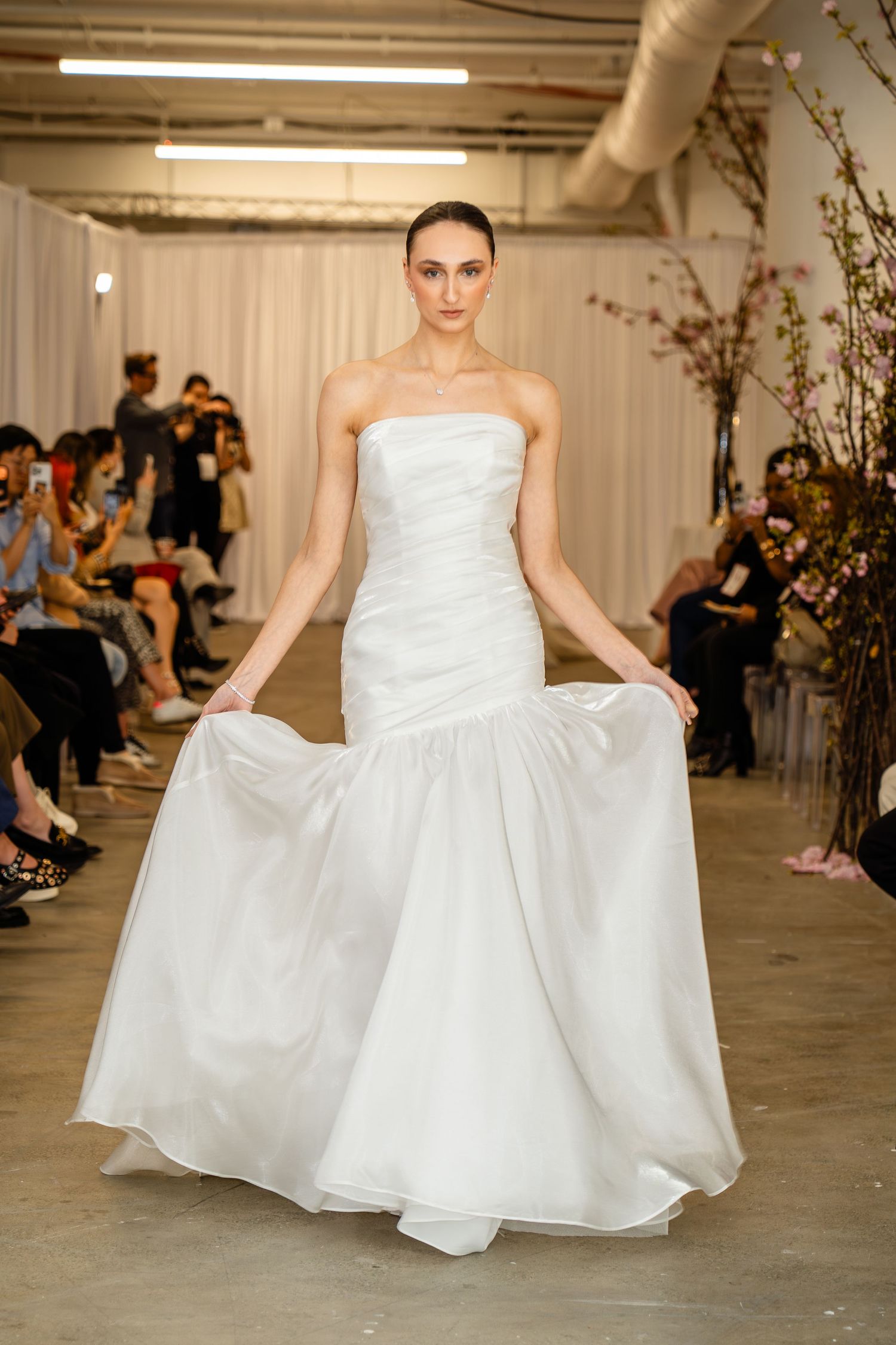 Model walks runway in strapless white mermaid wedding gown with flowing skirt at bridal fashion show.
