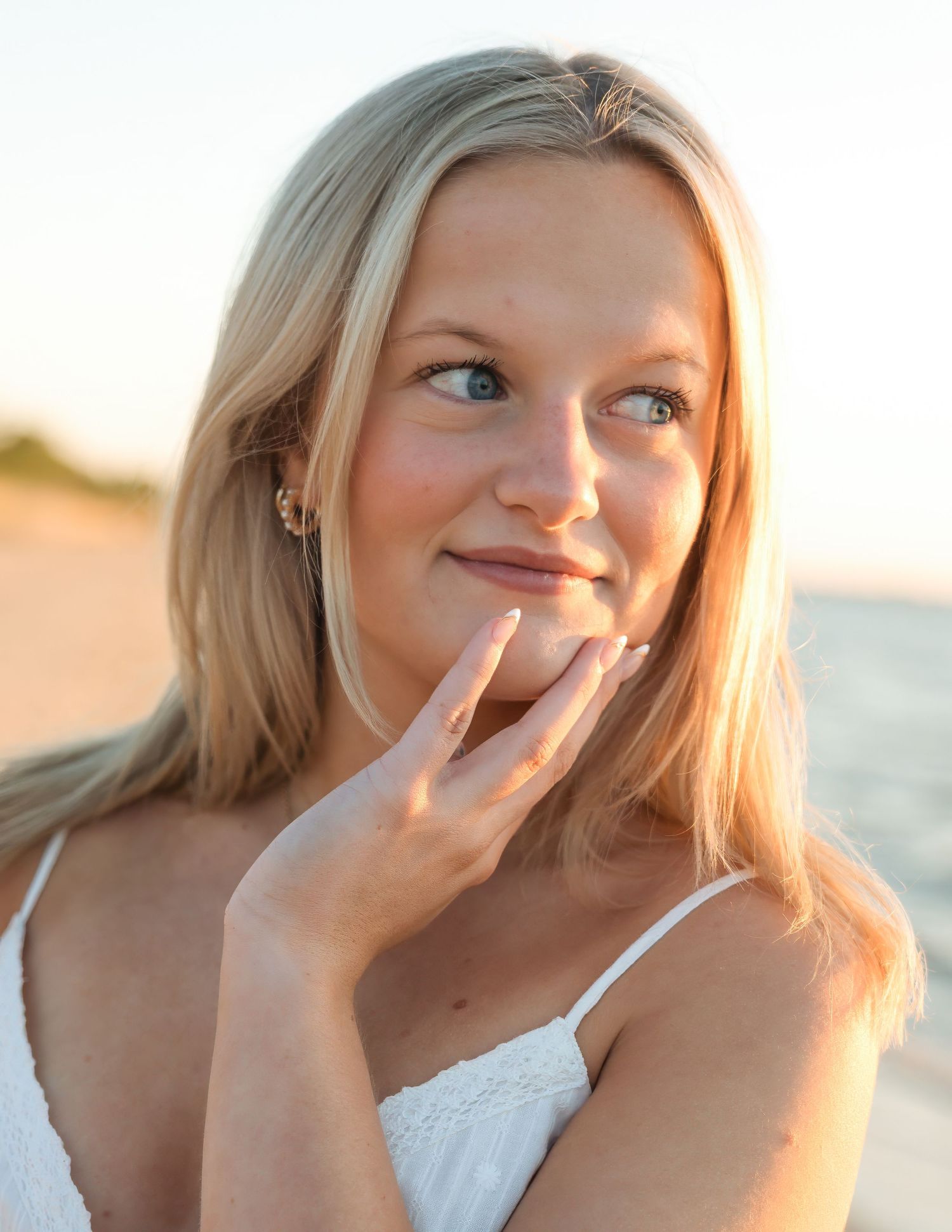 Smiling blonde woman with blue eyes poses on a sunny beach, wearing a white dress and touching her chin gently.