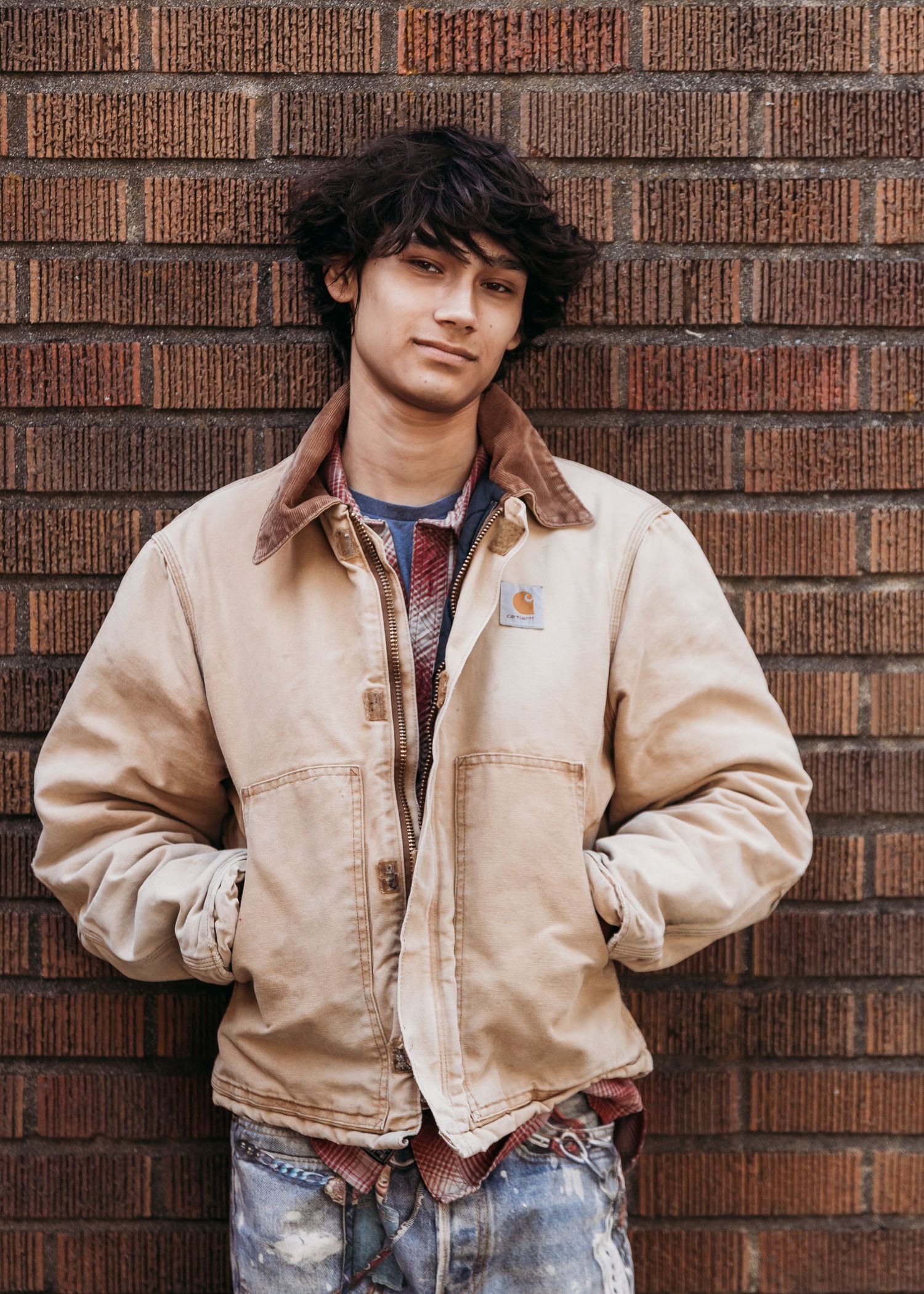 Young man in a tan Carhartt jacket leaning against a brick wall with hands in pockets.