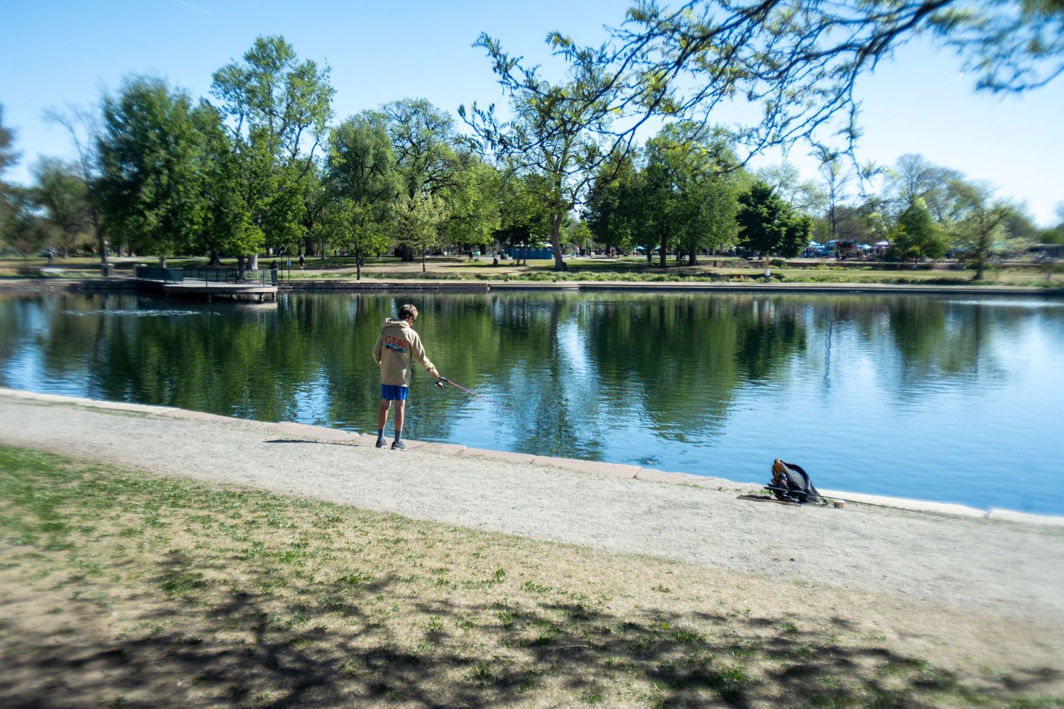 Person fishing at a calm urban park lake surrounded by lush green trees on a sunny day.