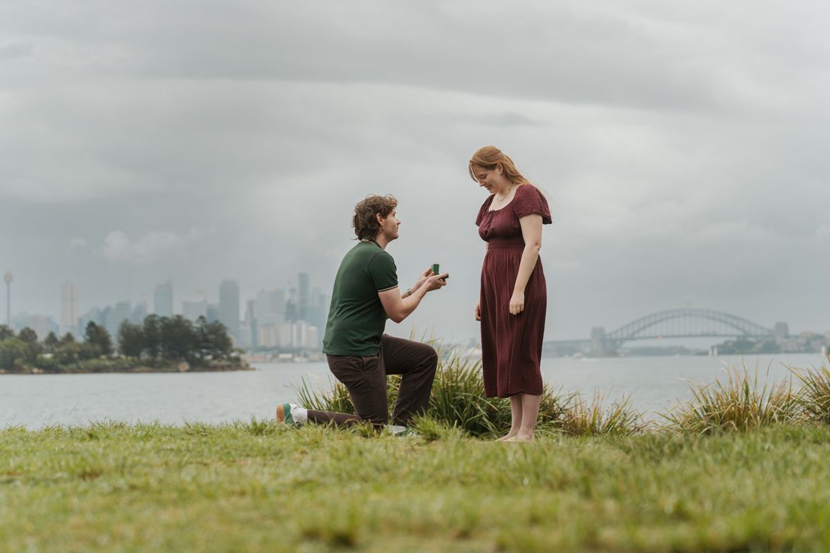 Man proposing to woman on grassy waterfront with Sydney city skyline and Harbour Bridge in background during a secret proposal photoshoot