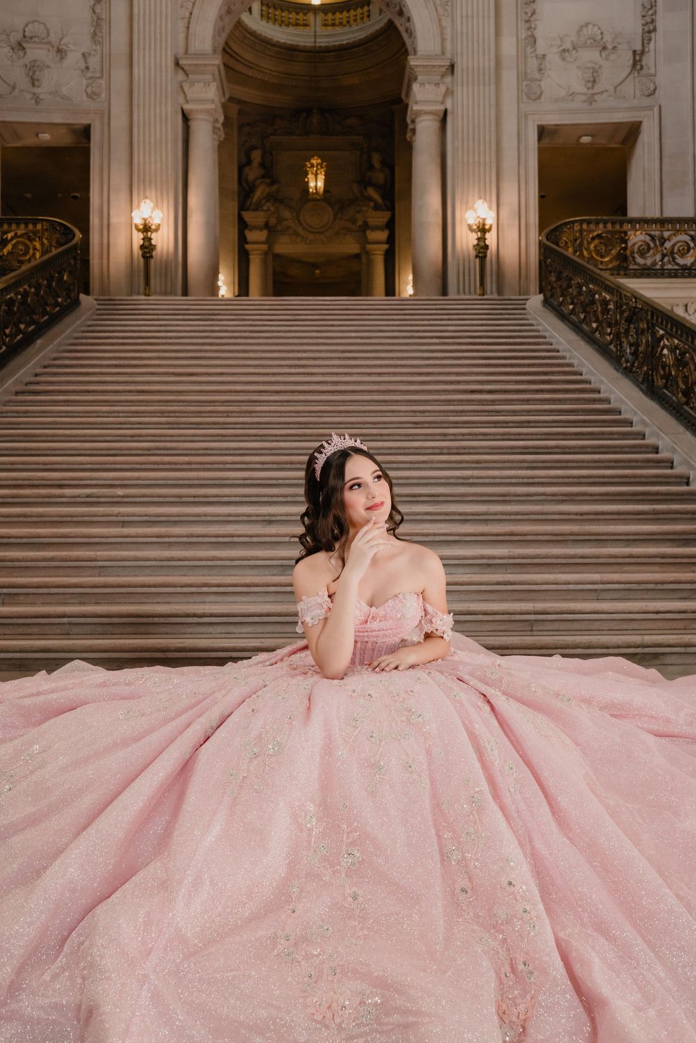 Young woman in pink ball gown quinceañera dress sits gracefully on grand marble staircase in elegant venue.