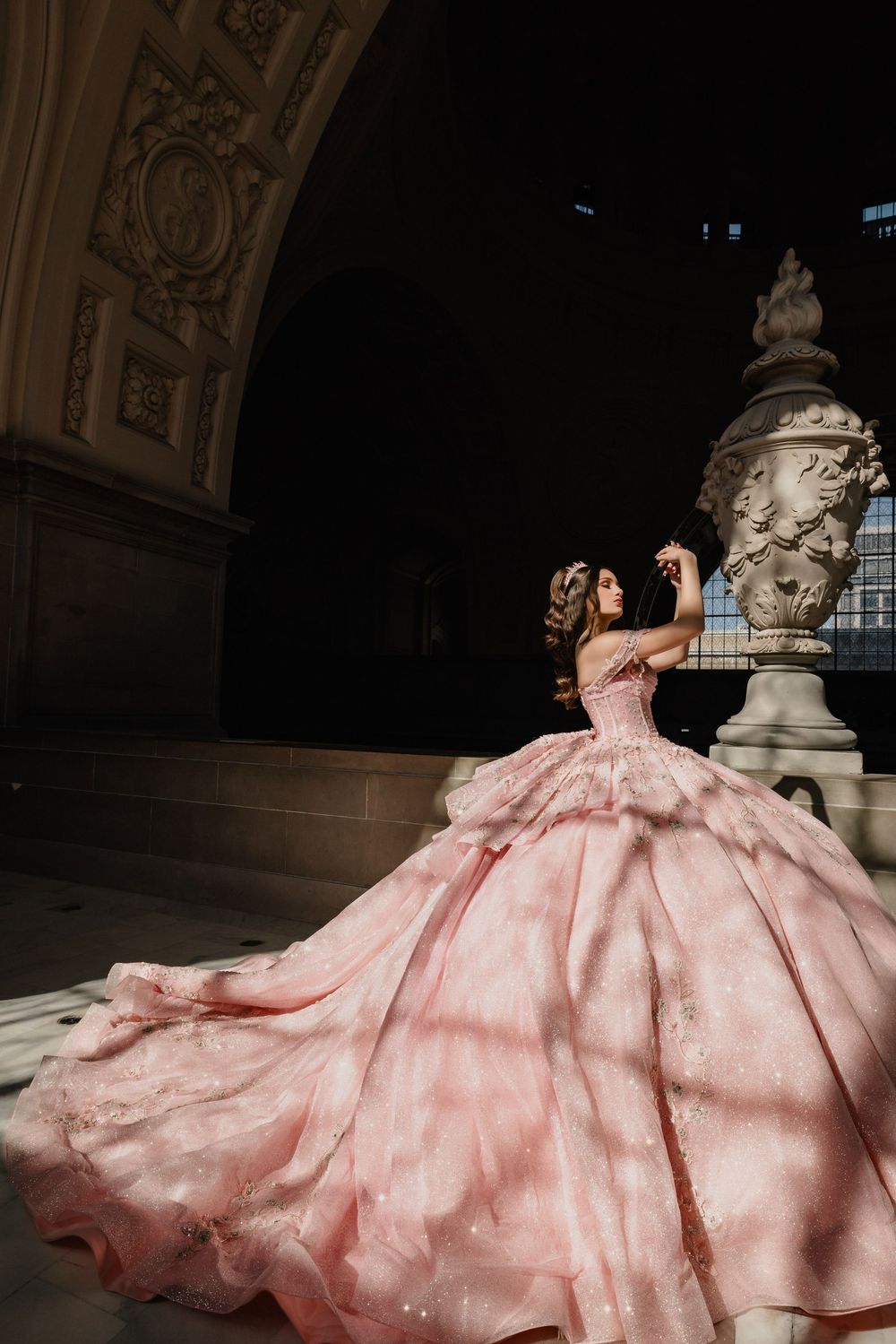 Woman in a stunning pink ball gown poses gracefully beside an ornate stone column in an elegant architectural setting.