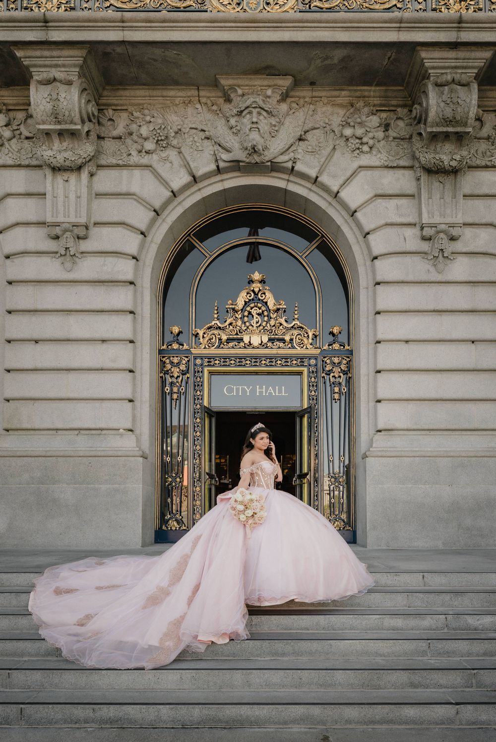 Bride in a flowing pink ball gown poses on City Hall steps beneath an ornate golden archway.