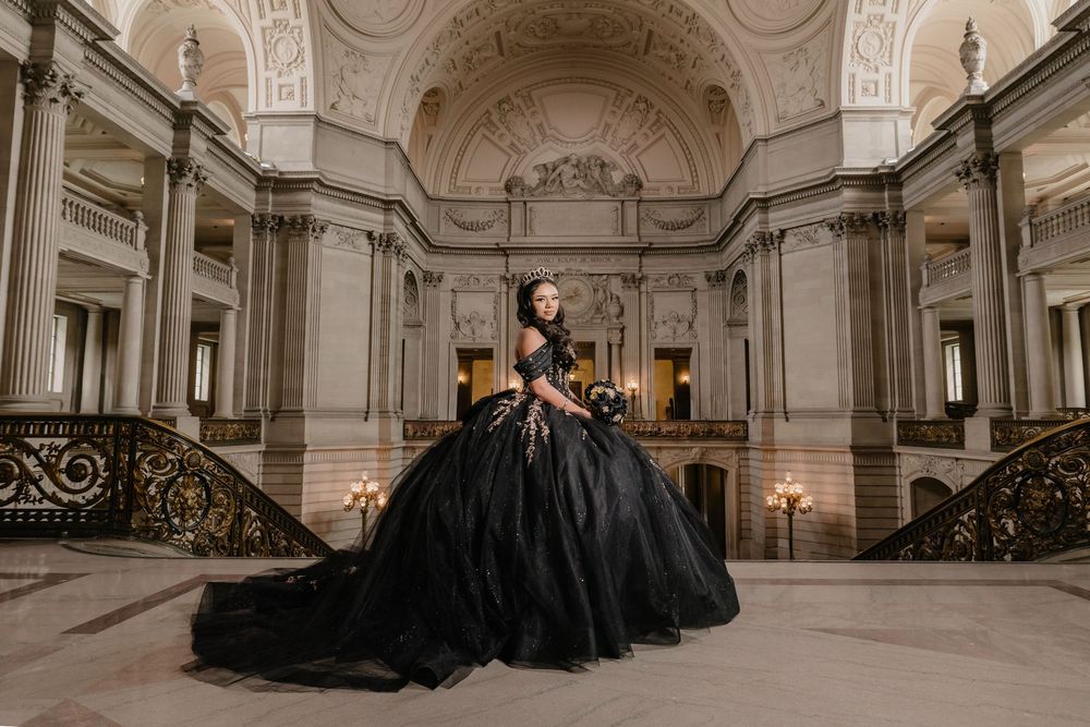 Woman in black ball gown poses elegantly in grand neoclassical hall with ornate staircases and columns.