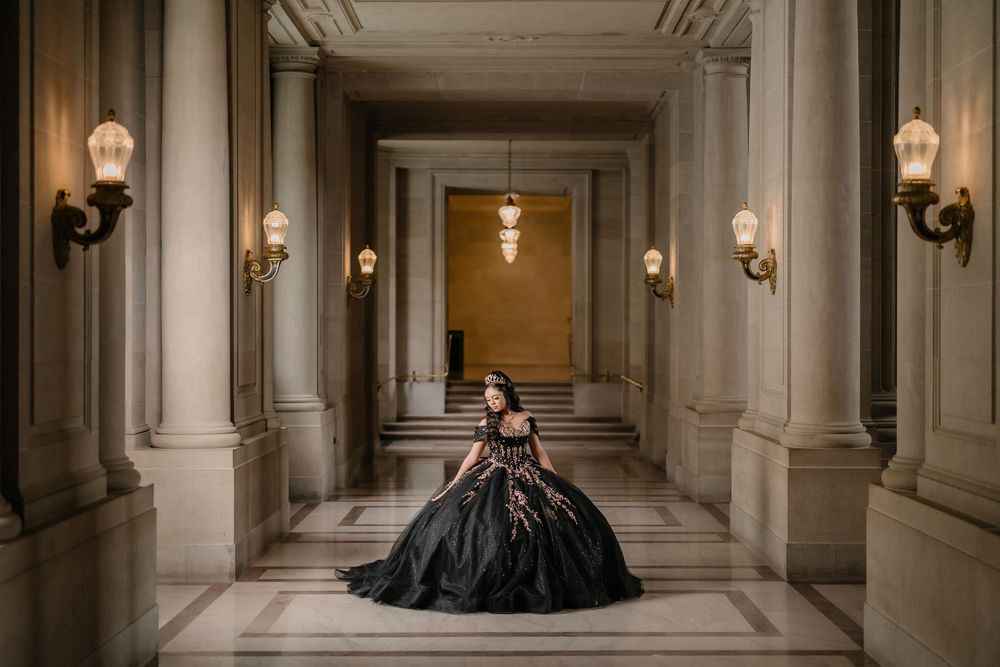 Woman in black ballgown poses in elegant marble hallway with ornate columns and warm wall sconces.